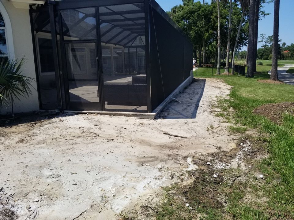 A backyard view showing a screened lanai enclosure alongside a patch of bare, sandy soil bordering a grassy lawn.