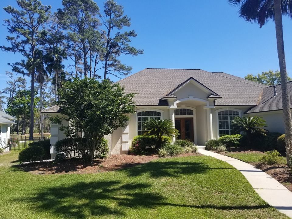A single-story, beige suburban home with a concrete walkway leading to the front door, surrounded by trees and lawn.