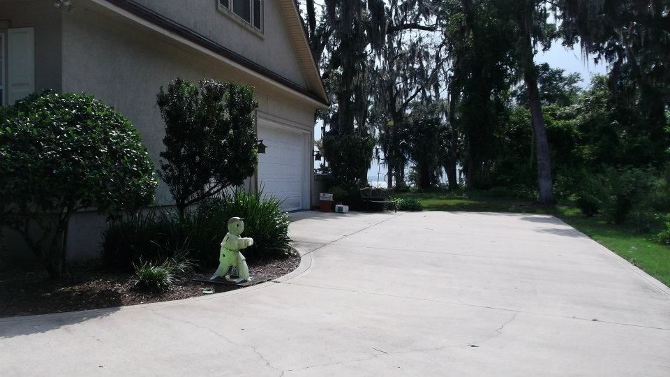 A concrete driveway leads to a suburban house garage next to a small, lime-green decorative statue in the garden.