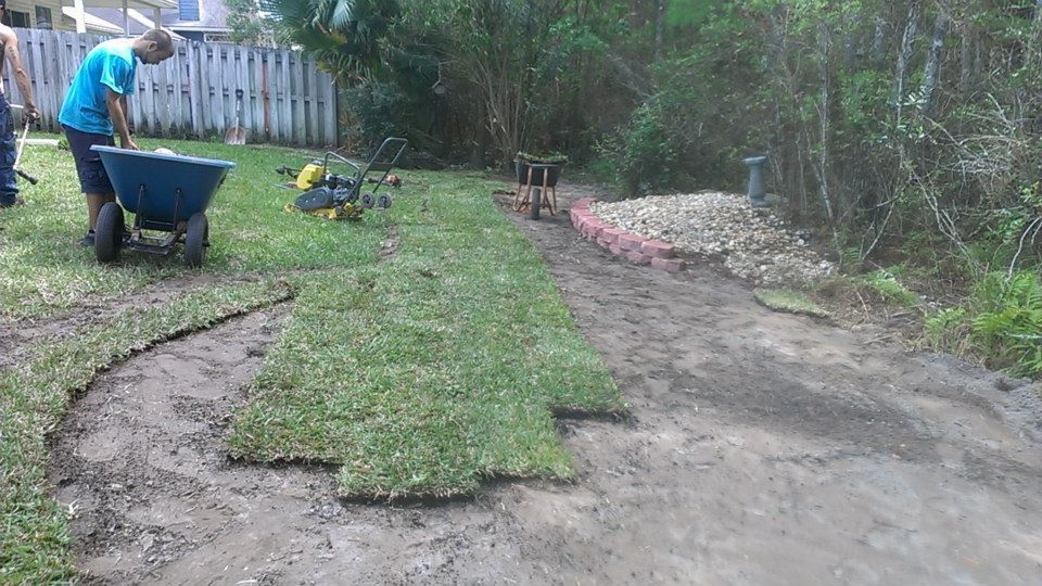 A person wearing a blue shirt uses a wheelbarrow while laying new sod in a backyard near a rock-bordered flower bed.