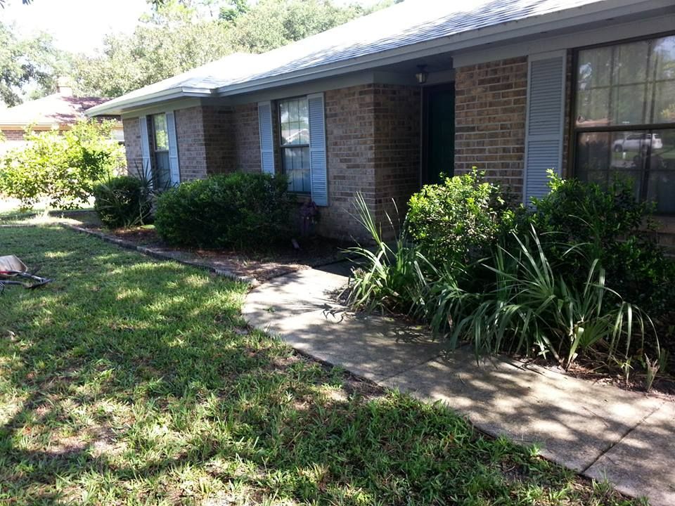 A brick house with a concrete walkway leading to the front door, surrounded by green shrubs and a grassy lawn.