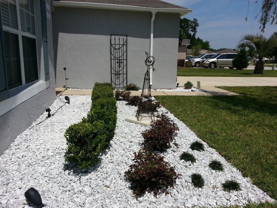 A landscaped front yard garden bed featuring white rock, trimmed green shrubs, reddish bushes, and a metal lawn ornament.