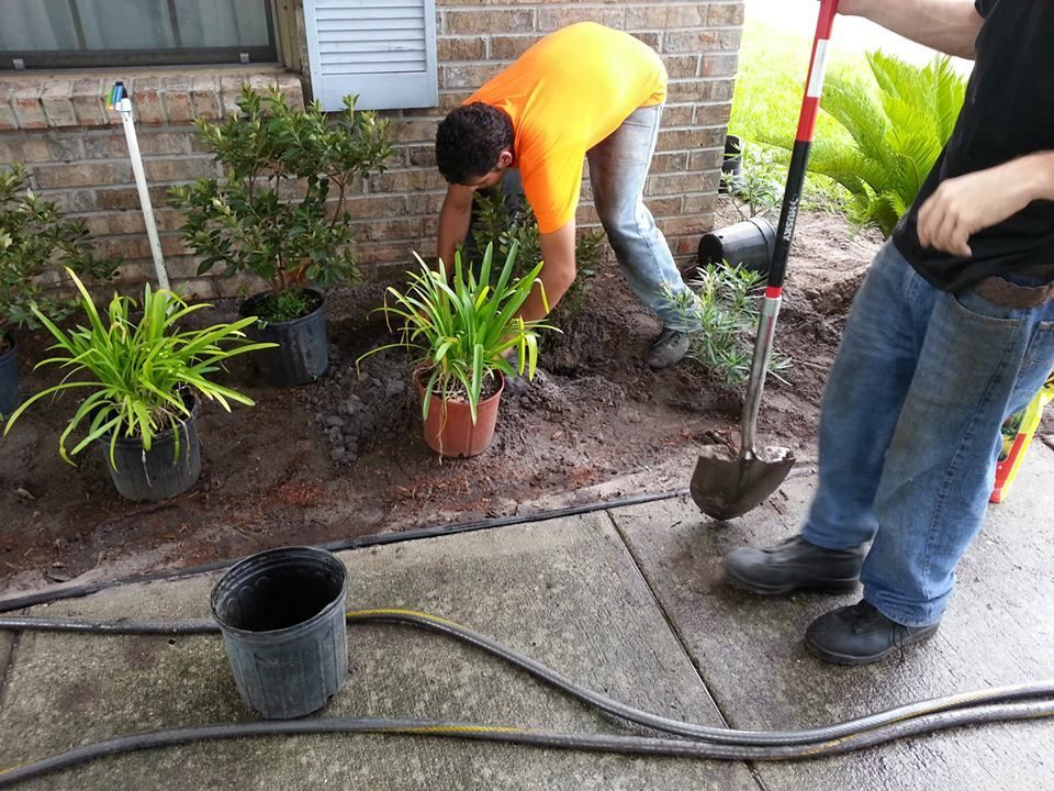 Two people plant flowers in a garden bed near a brick wall and sidewalk, one using a shovel.