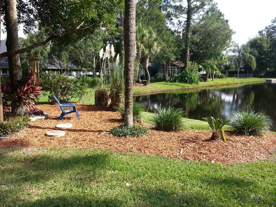 A blue Adirondack chair sits on a mulched garden bed beside a pond, surrounded by trees and decorative grasses.