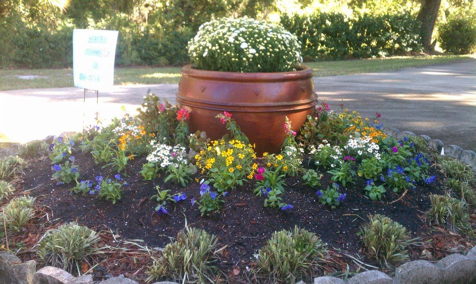 Flower bed with a large terracotta pot centerpiece and colorful blooms in a roadside garden.