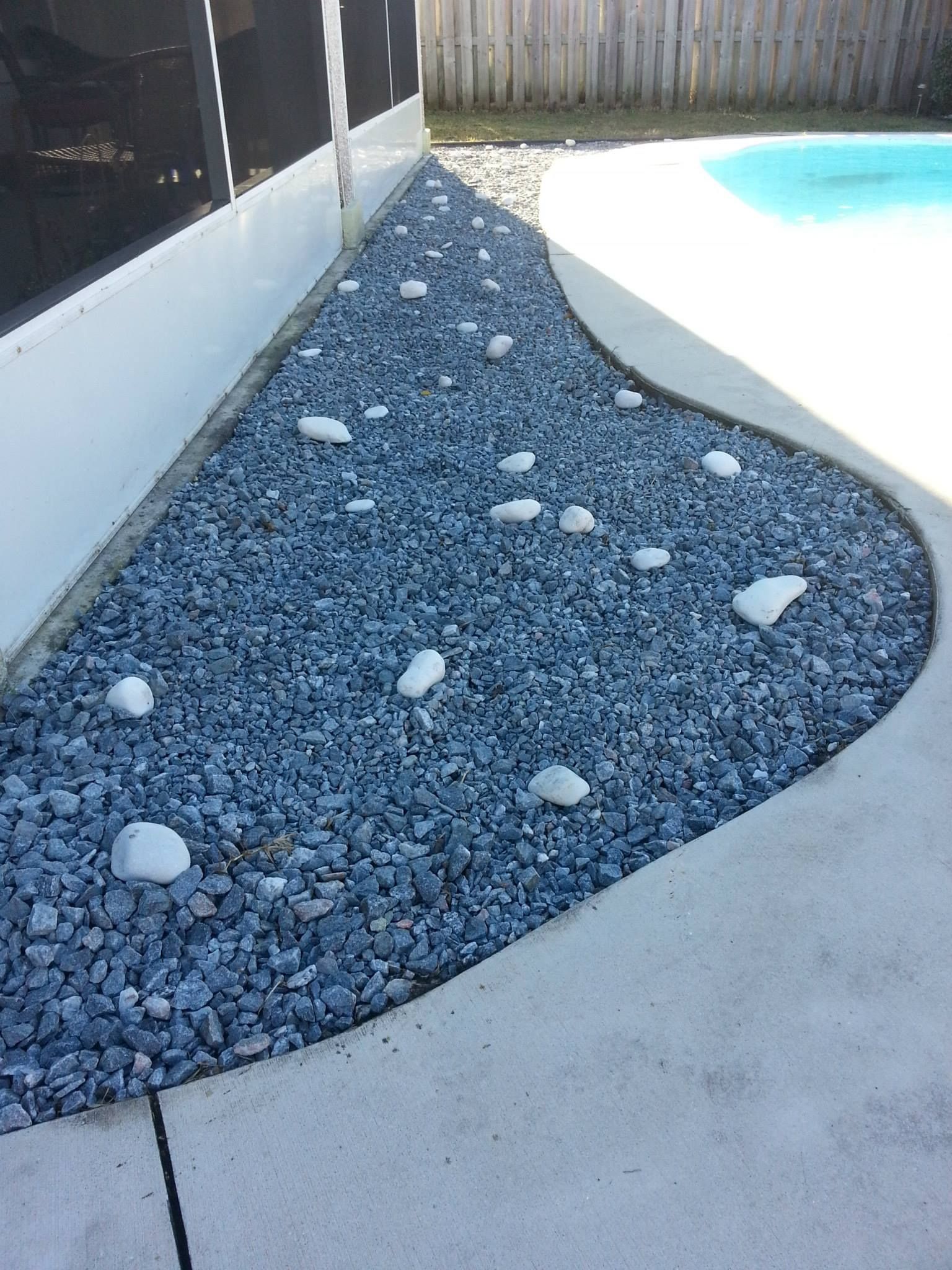 A narrow landscaped area beside a pool, filled with dark grey gravel and scattered white decorative stones.