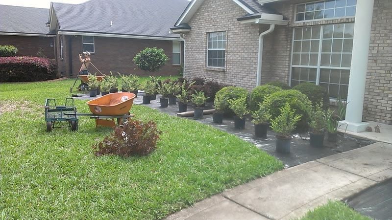 A person works on landscaping in front of a brick house, arranging numerous potted plants on a black garden fabric base.