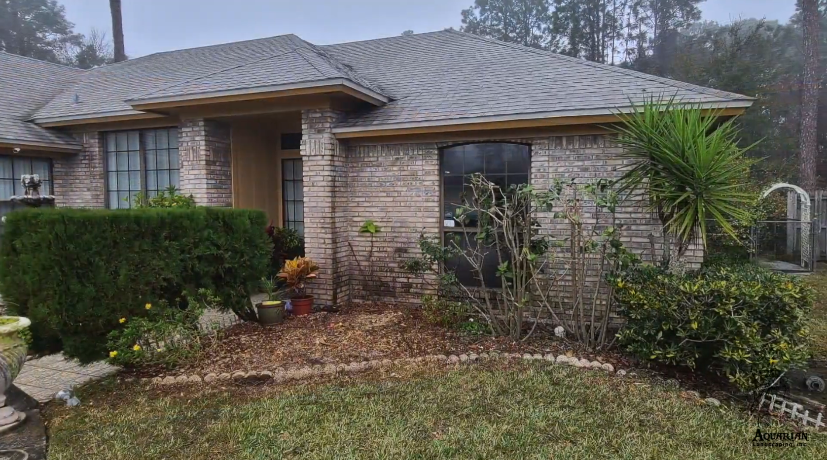 Single-story brick house with a front porch, large windows, shrubs, and a wet yard on a cloudy day