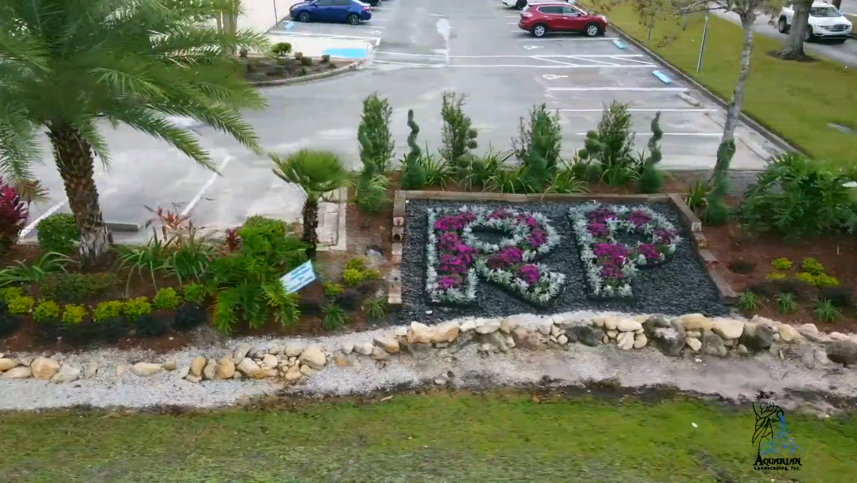 Letter “RP” made from flowers in a landscaped garden beside a parking lot.