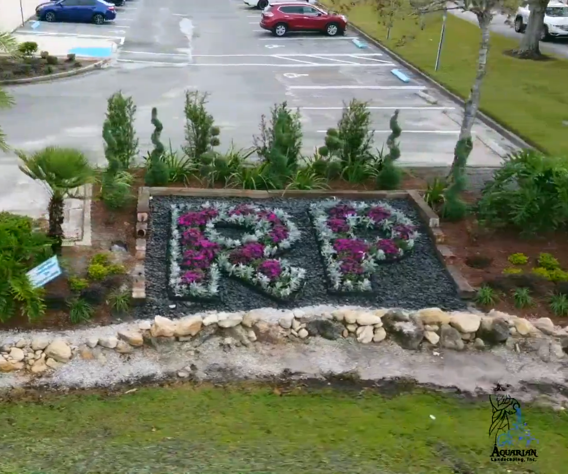 Flower bed spelling “RP” beside a parking lot, bordered by rocks and shrubs.