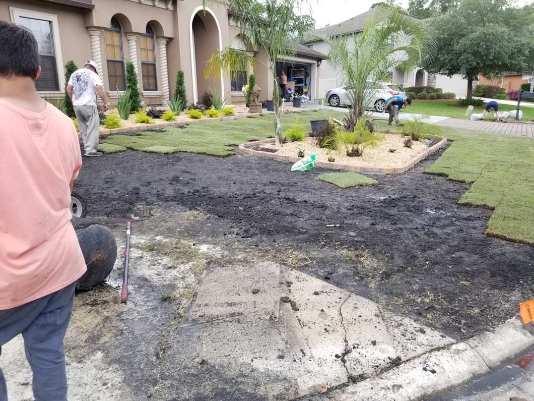 Two people landscape a suburban yard, installing new sod patches over freshly prepared soil near a concrete driveway.