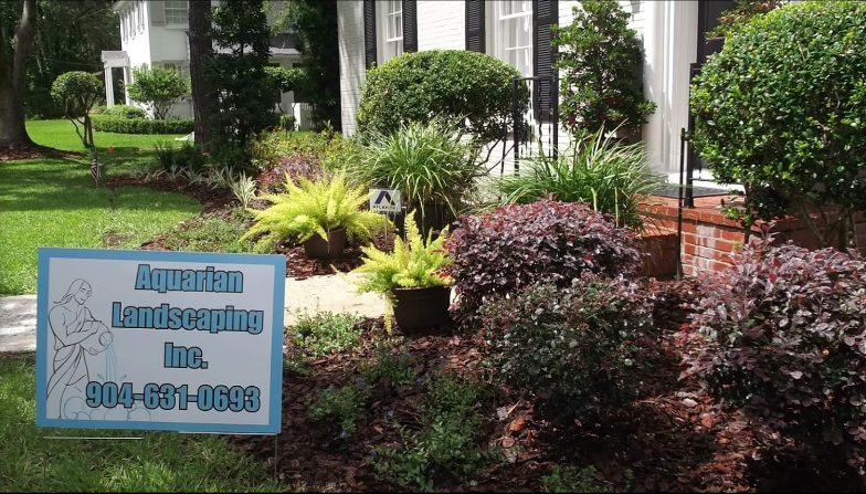 A sign for Aquarian Landscaping Inc. with a phone number, placed in front of a house with lush, manicured garden beds.