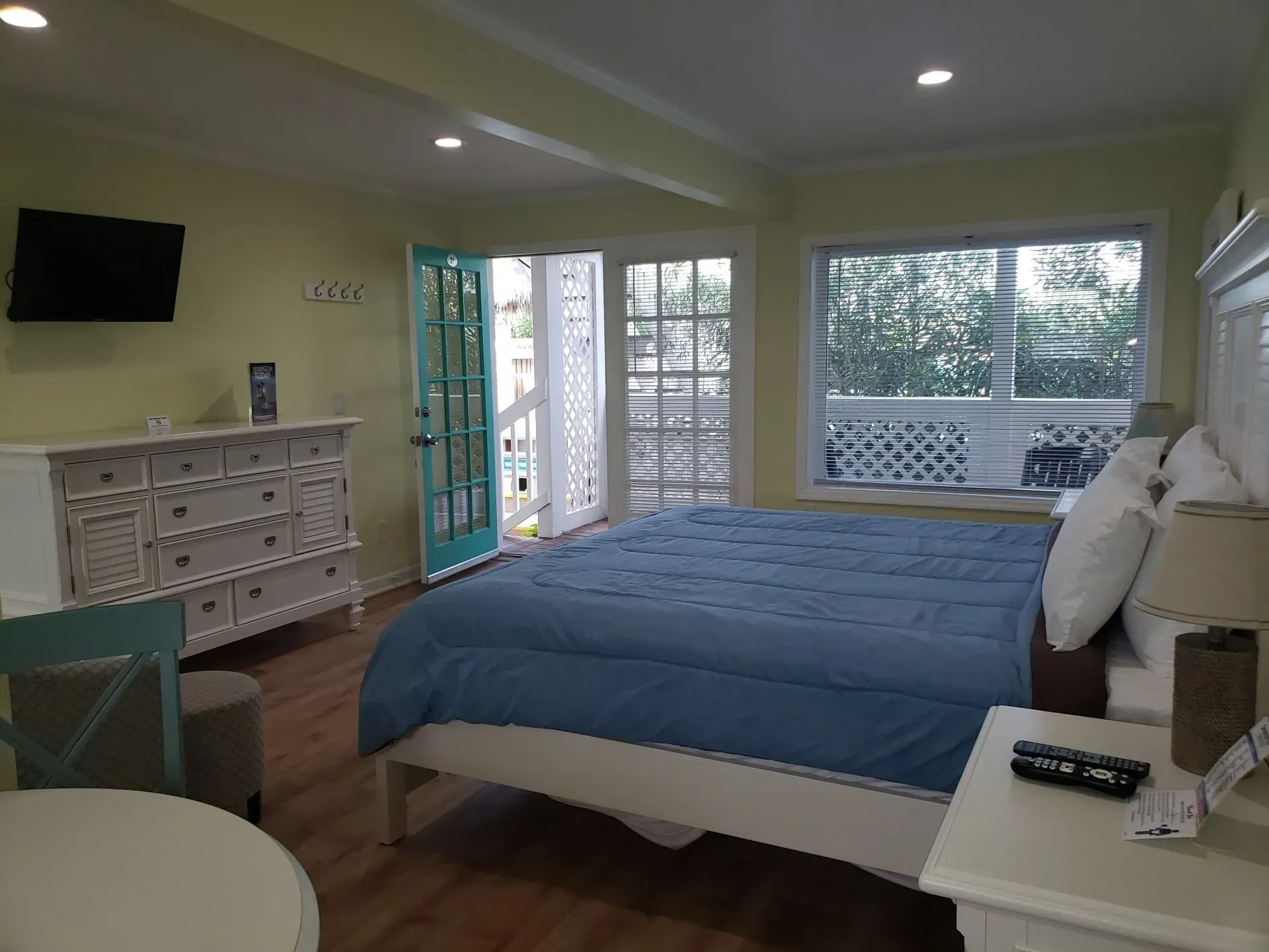 A bedroom featuring a bed with a blue comforter, white dresser, light-colored walls, and a teal door leading outside.