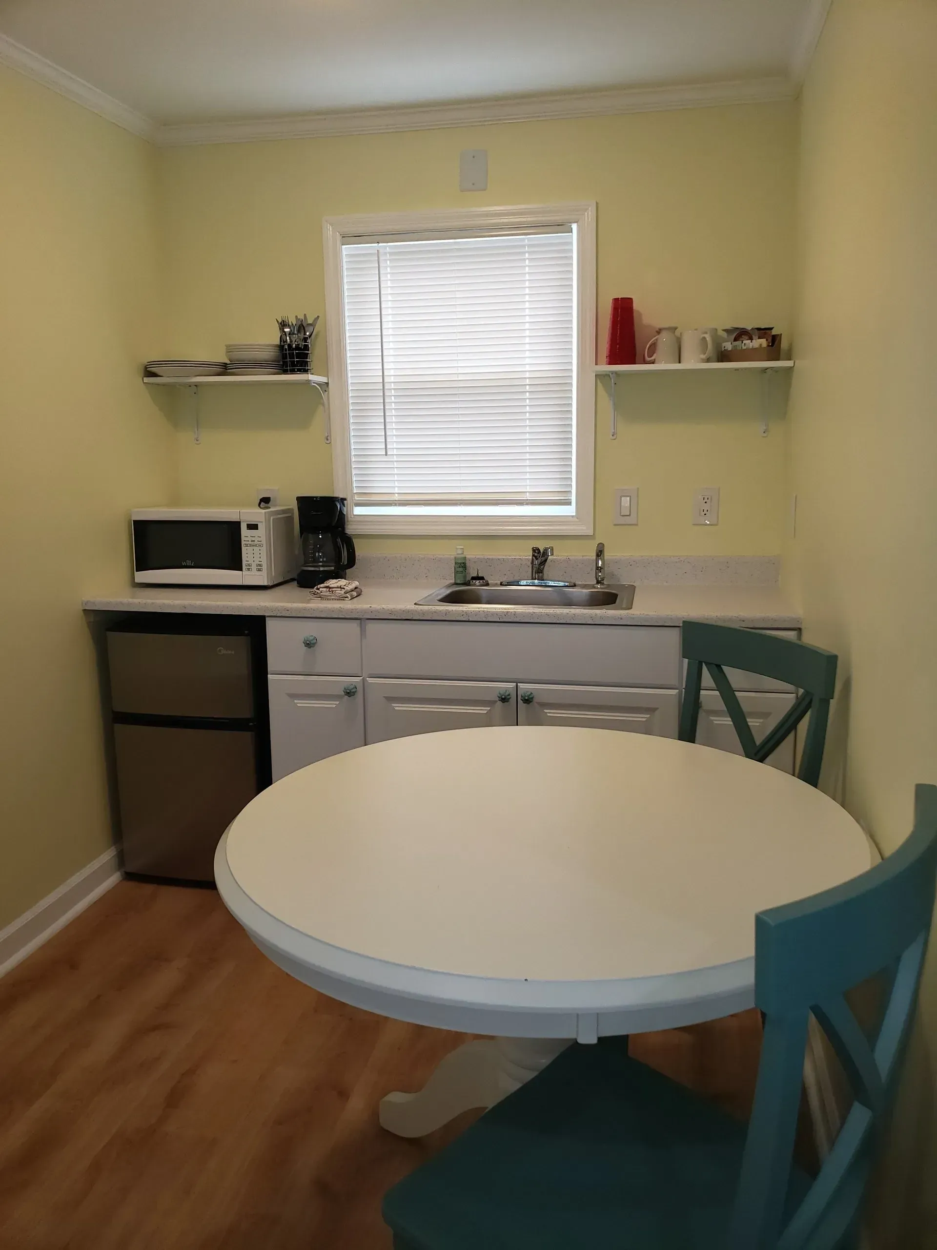 A small kitchen featuring pale yellow walls, white cabinets, a microwave, sink, mini fridge, and a white table with a chair.