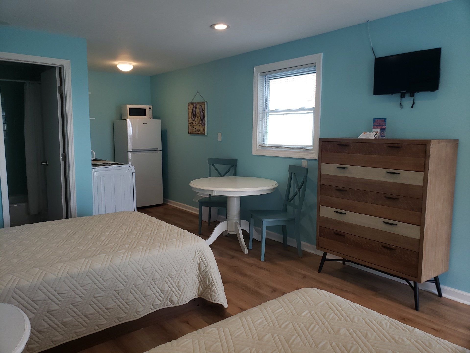 A hotel room with light blue walls, two beds, a round white table with two chairs, a refrigerator, and a dresser.