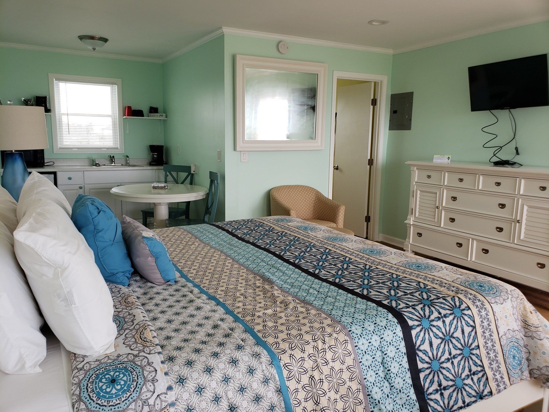 A bright bedroom with pale green walls, a patterned bedspread, blue accent pillows, a white dresser, and a small dining set.