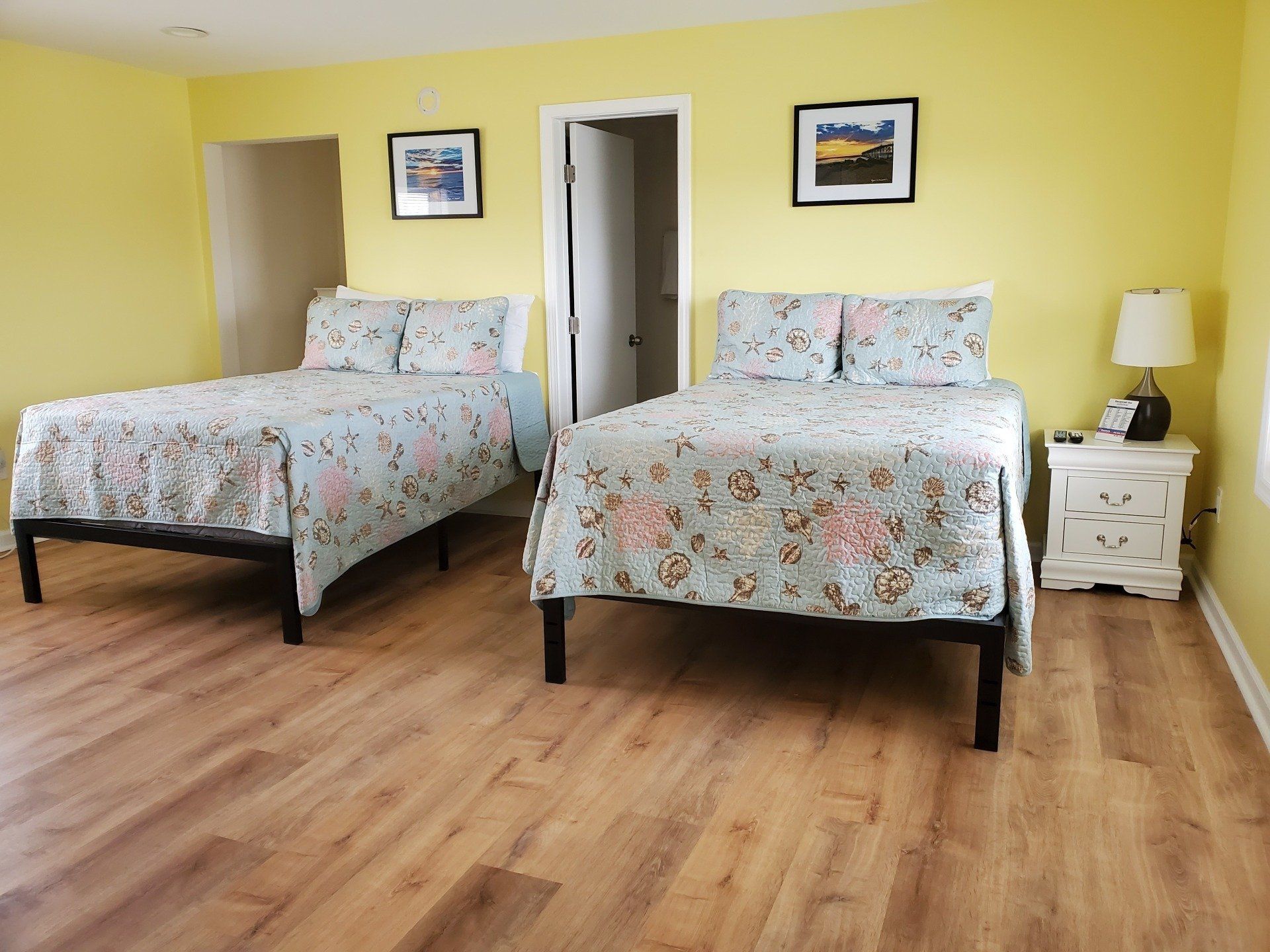 Two beds with floral patterned quilts in a room with light yellow walls and wood-look flooring, featuring two framed prints.