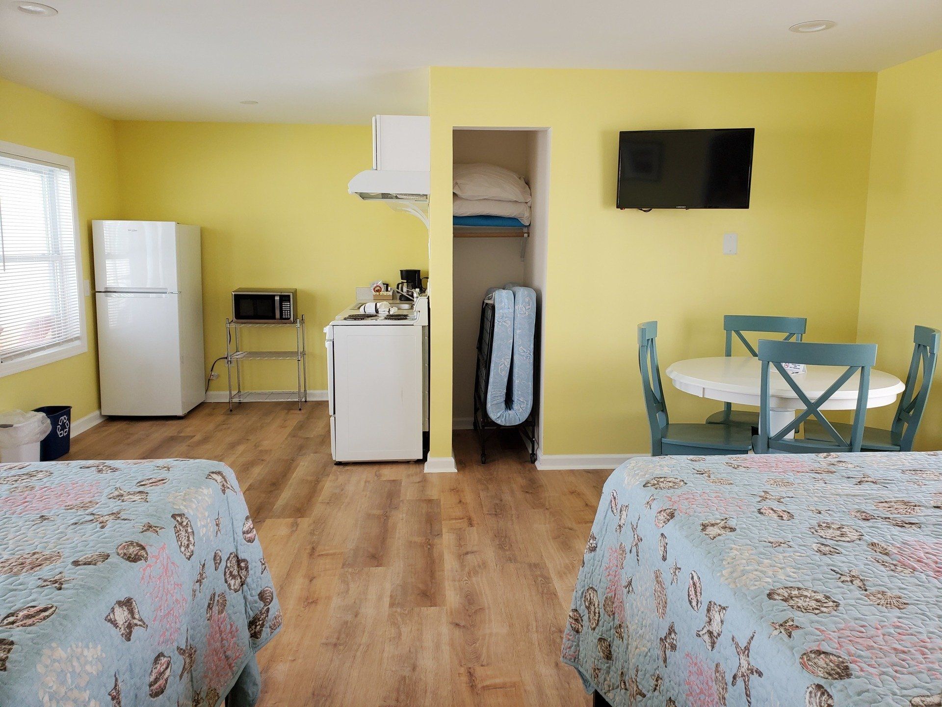 A yellow-walled motel room with two patterned beds, a small kitchen area with a fridge, and a dining table for four.