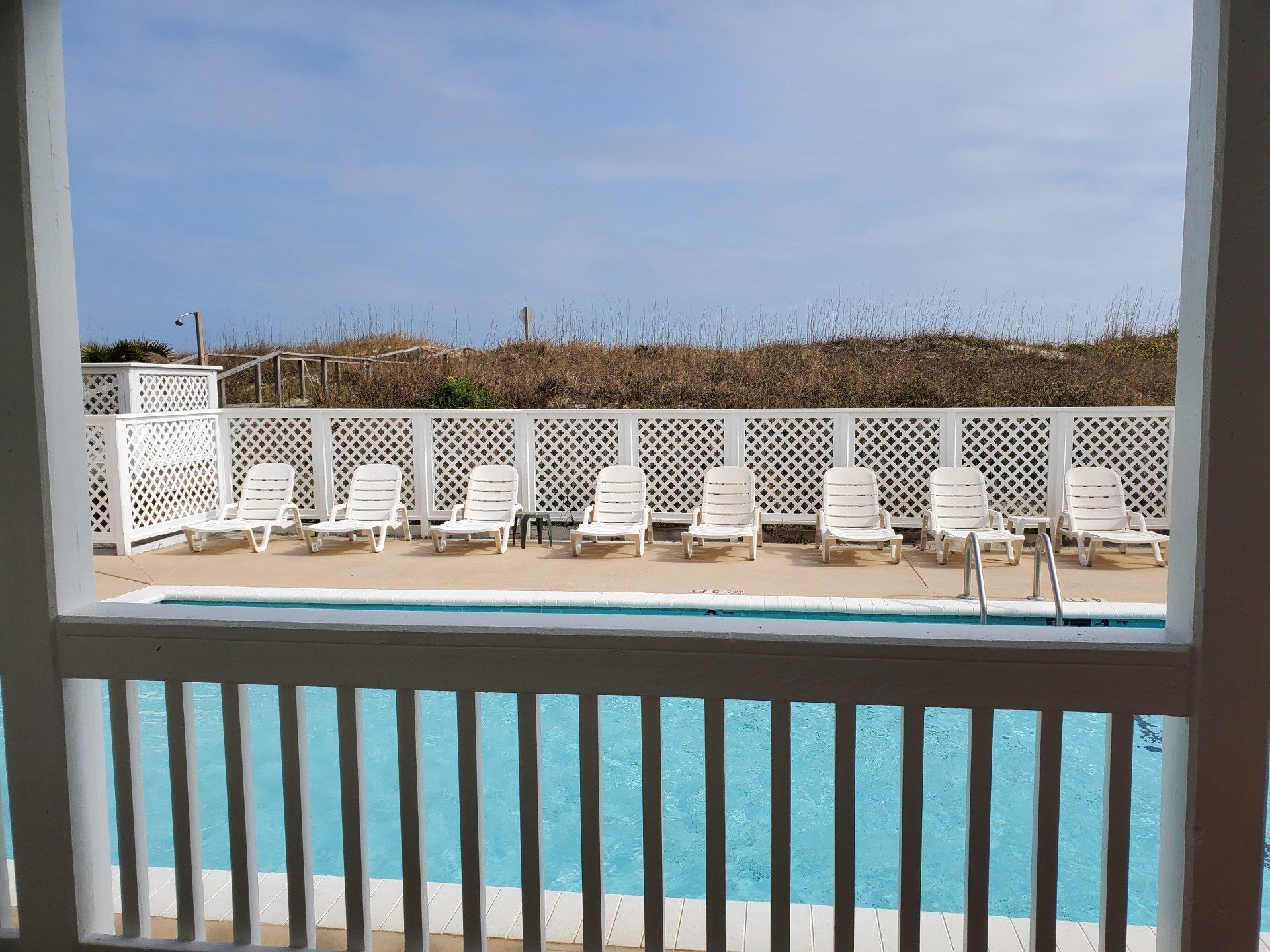 A swimming pool with lounge chairs behind a white railing, overlooking sand dunes and a clear sky.