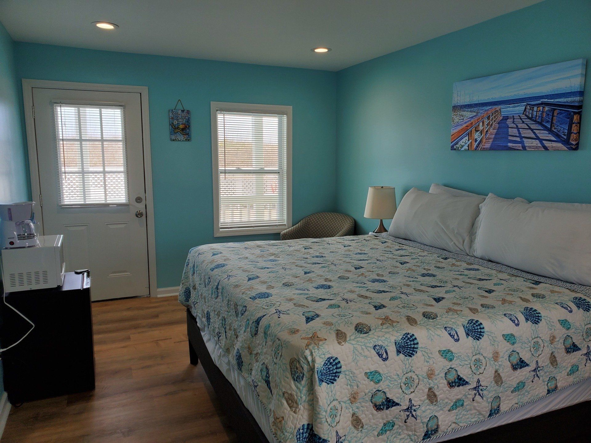A bedroom with light blue walls, a bed with a seashell-patterned quilt, a door, a window, and a coastal-themed wall painting.