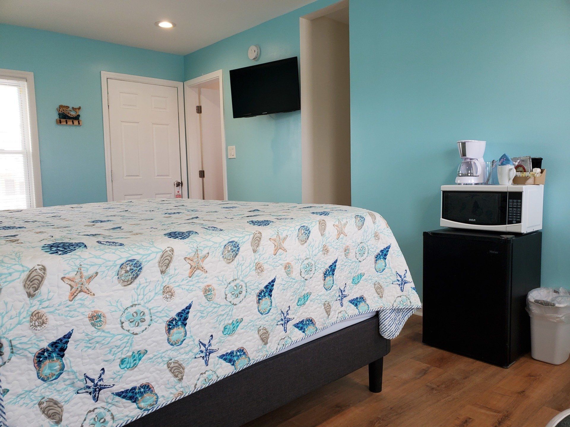 A bright bedroom with teal walls, a bed with a seashell-patterned quilt, a wall-mounted TV, and a mini-fridge station.
