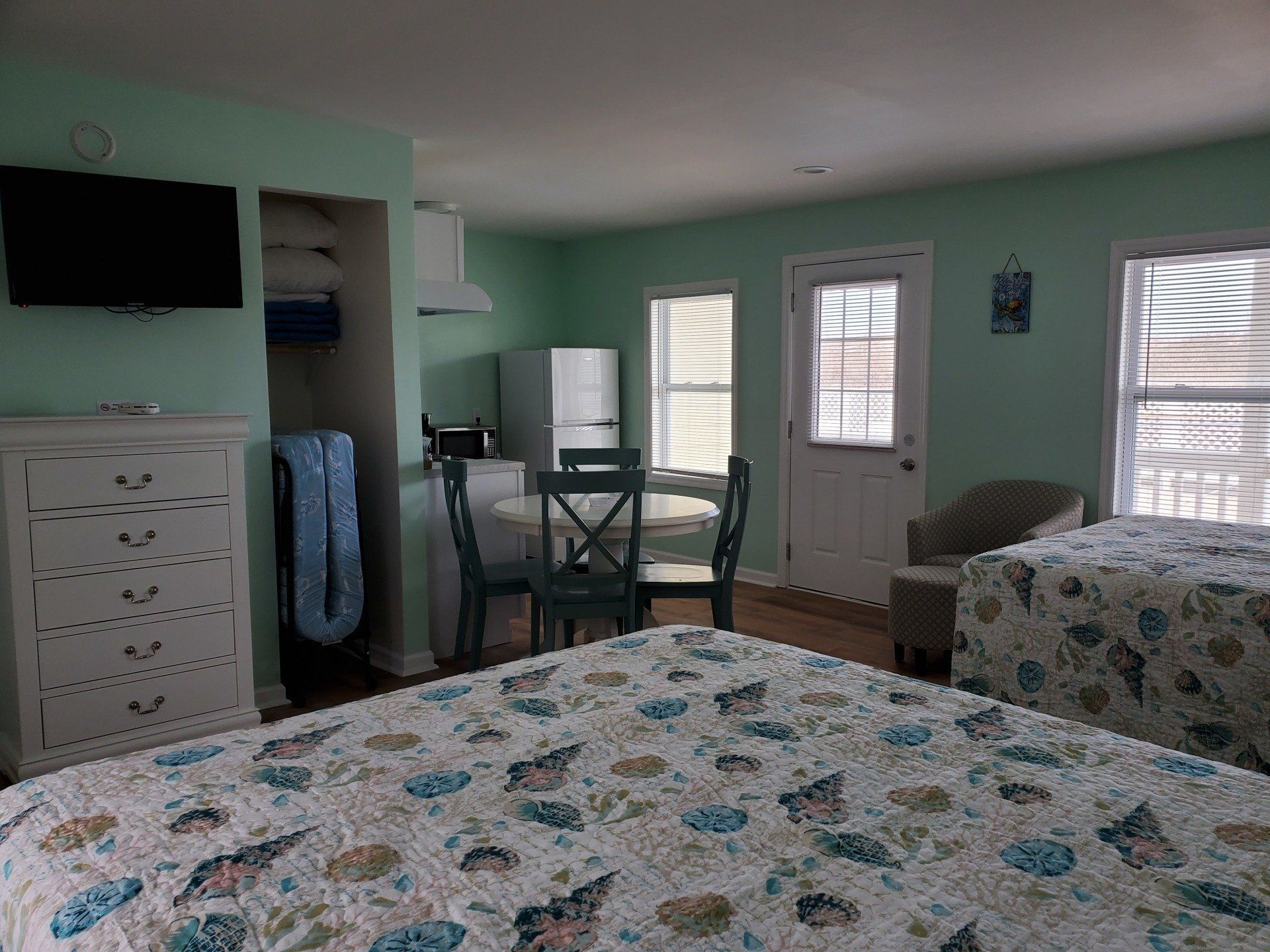 A light green hotel room with two beds featuring seashell quilts, a white dresser, and a small dining table.