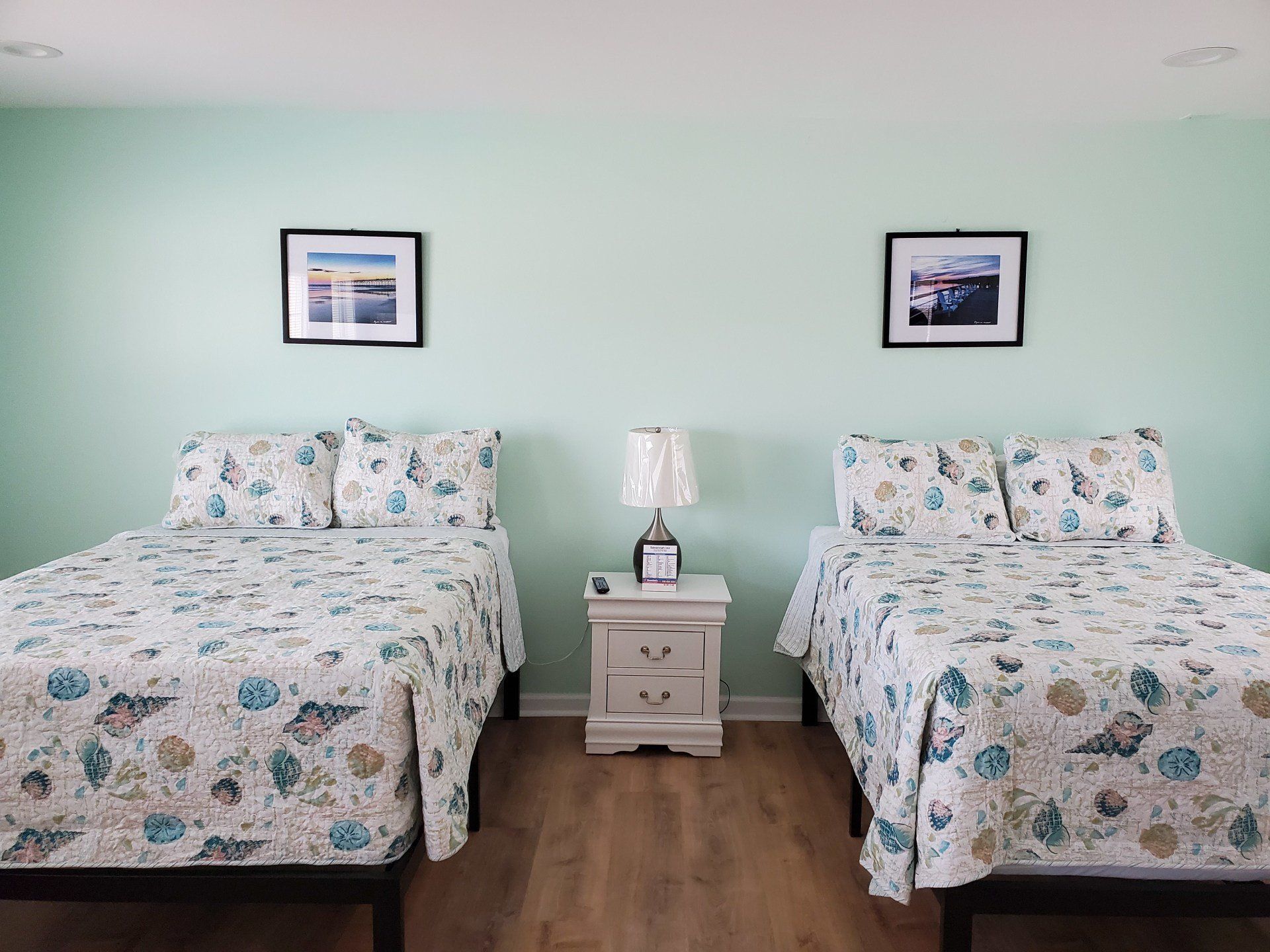 Two beds with seashell-patterned bedding sit in a light green room with a white nightstand and two framed wall pictures.