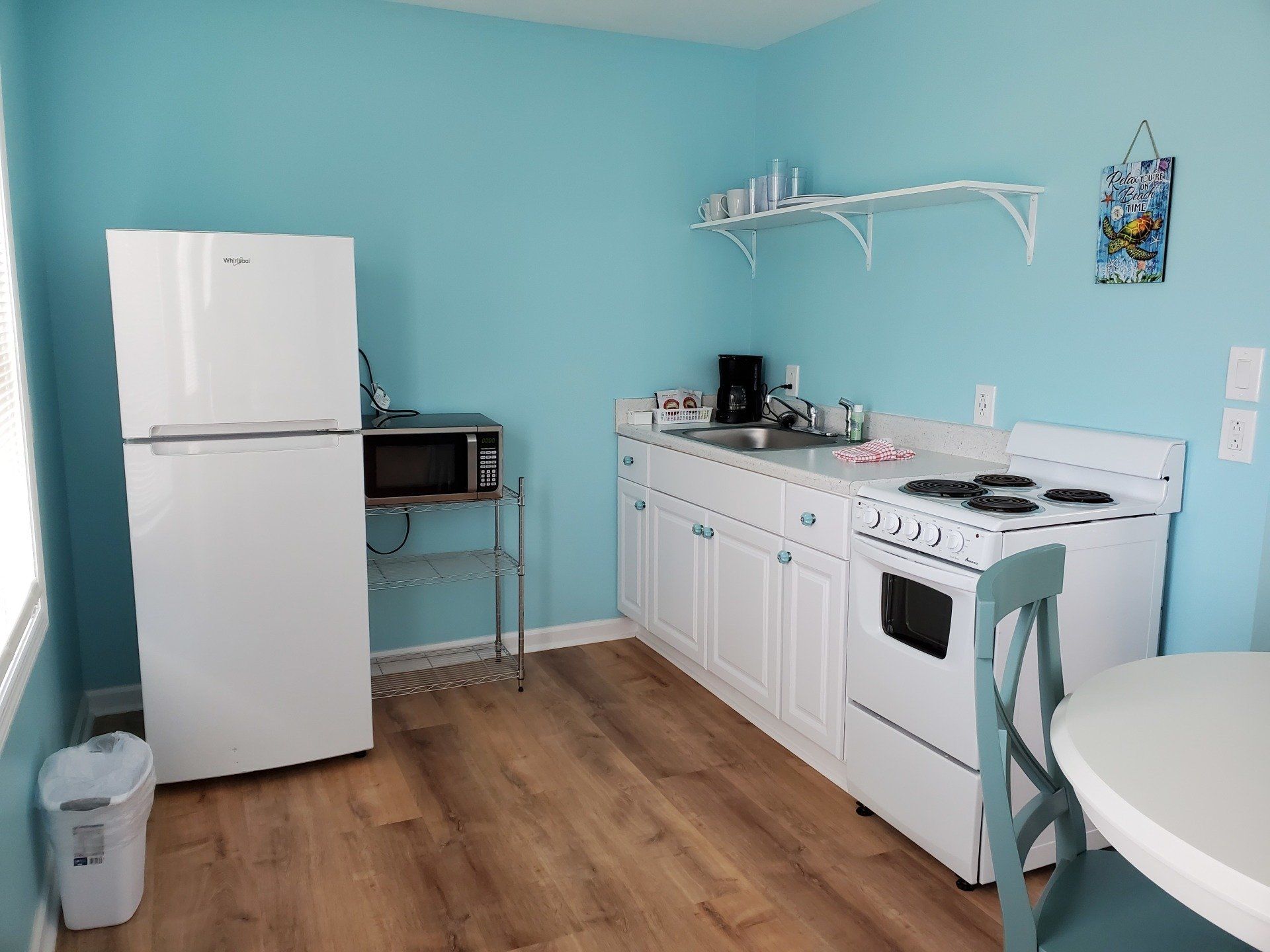 A small kitchen with light blue walls, wood floors, a white refrigerator, stove, microwave, and a wooden chair.