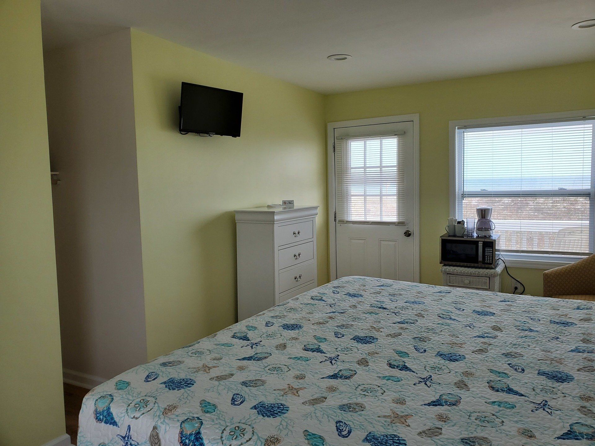 A bedroom with pale yellow walls, a bed with a seashell-patterned quilt, a white dresser, a television, and a window.