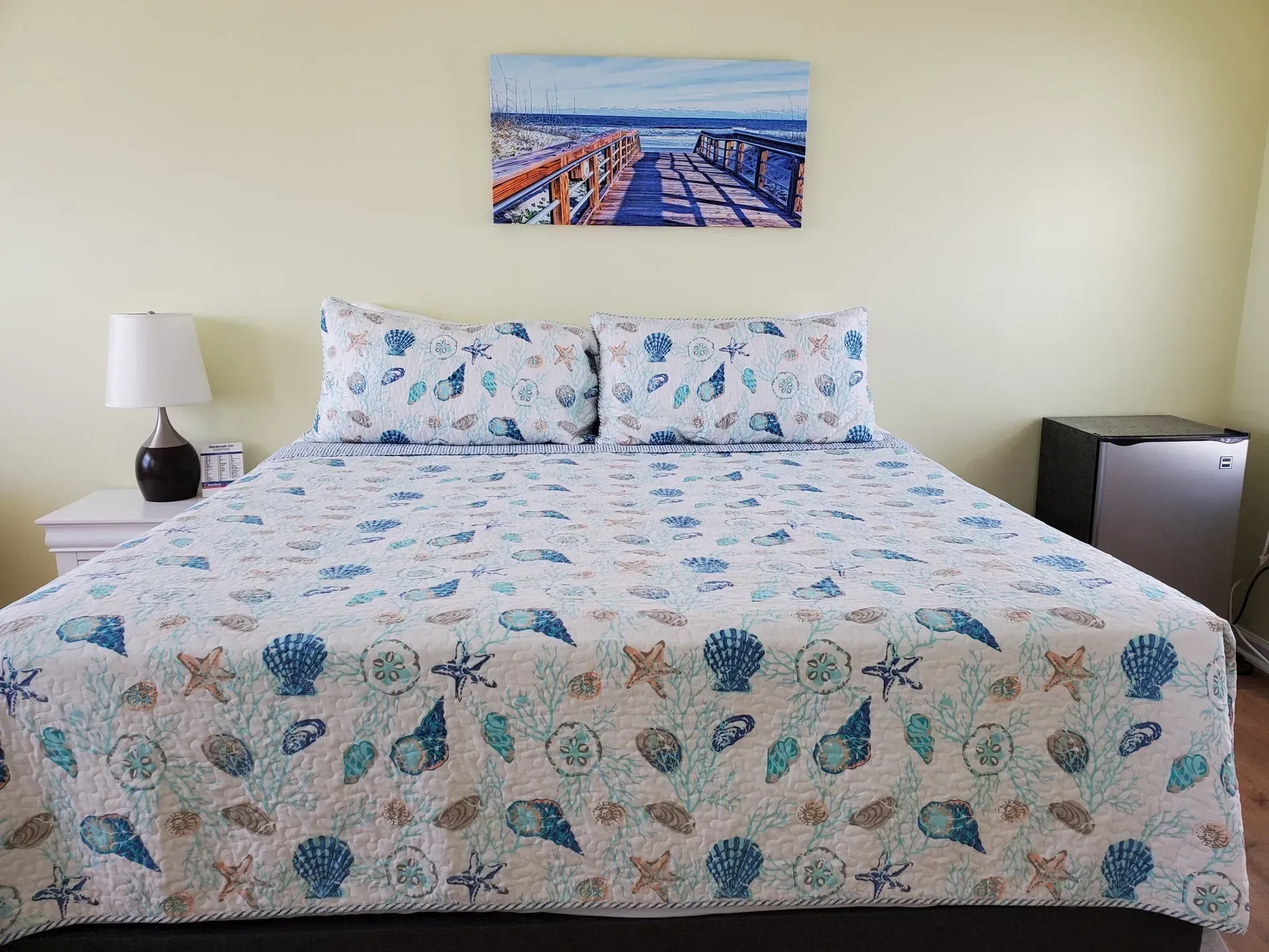 A neatly made bed with a blue and white seashell-patterned quilt, a coastal print above, and a lamp on the side table.