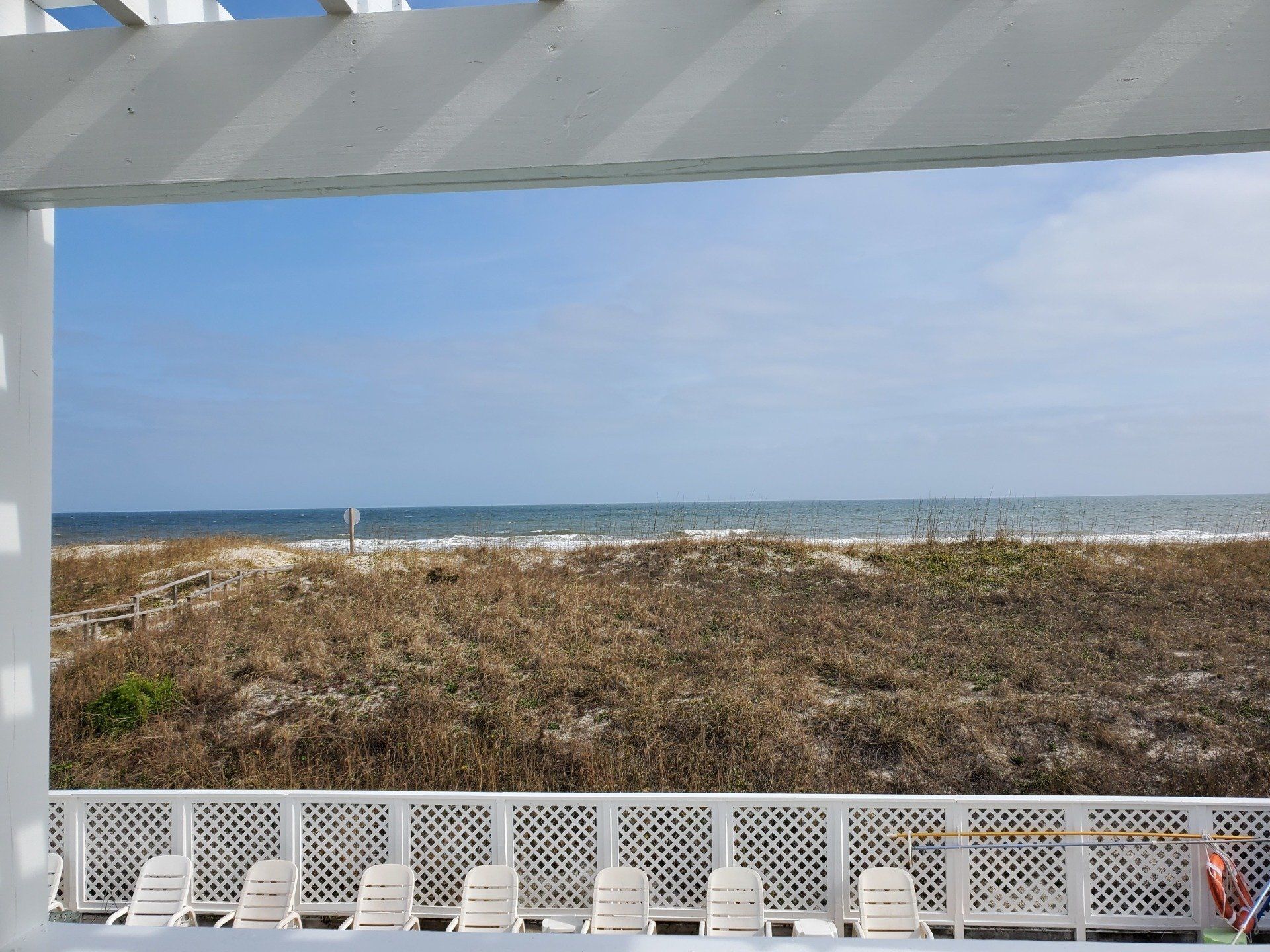 A white lattice railing and chairs overlook sand dunes and the ocean under a clear sky.