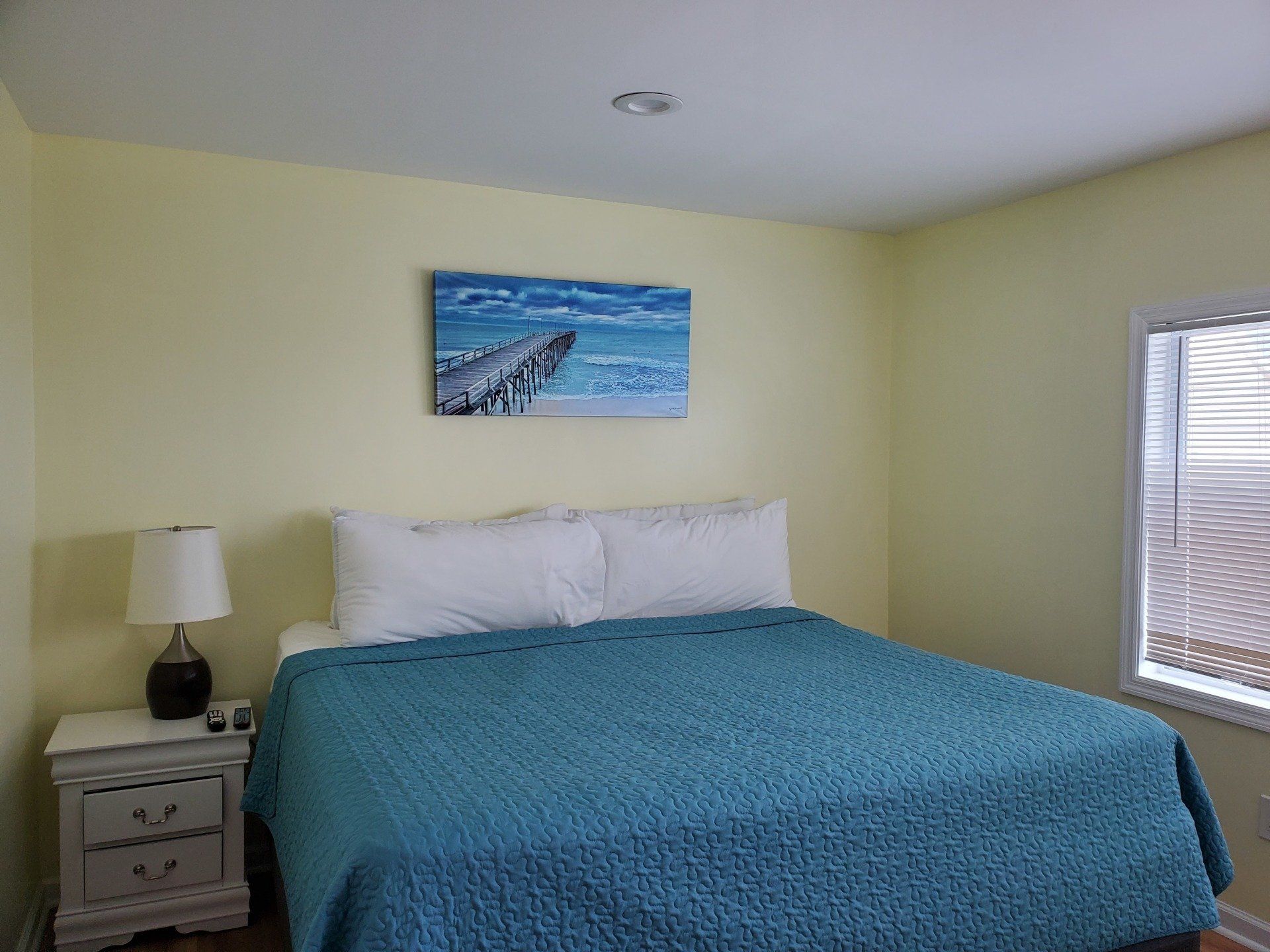 A bedroom with a king-sized bed, teal textured quilt, white pillows, nightstand with a lamp, and a beach wall painting.