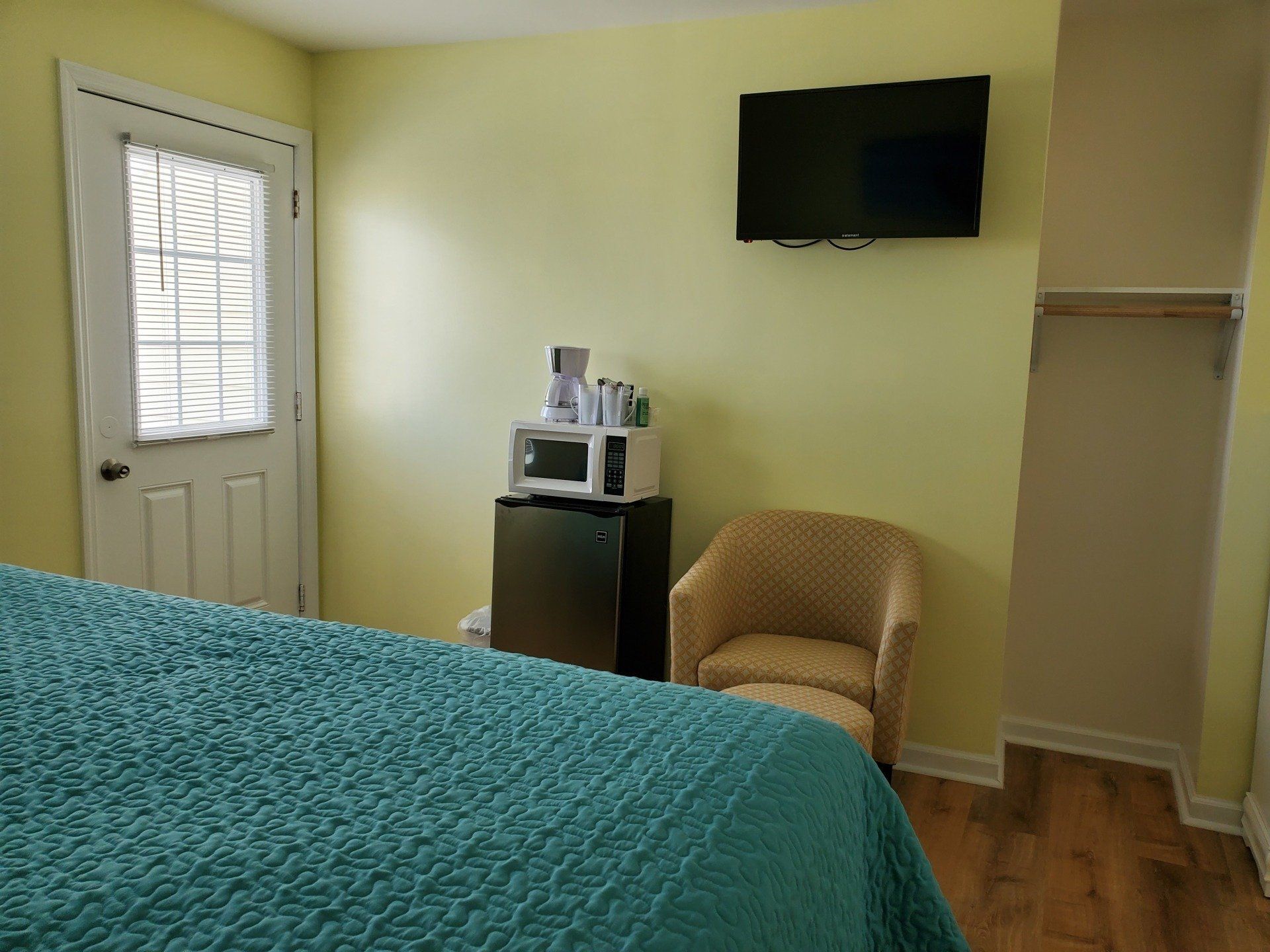 A hotel room with a teal bed, light yellow walls, a small microwave on a mini-fridge, an armchair, and a wall-mounted TV.