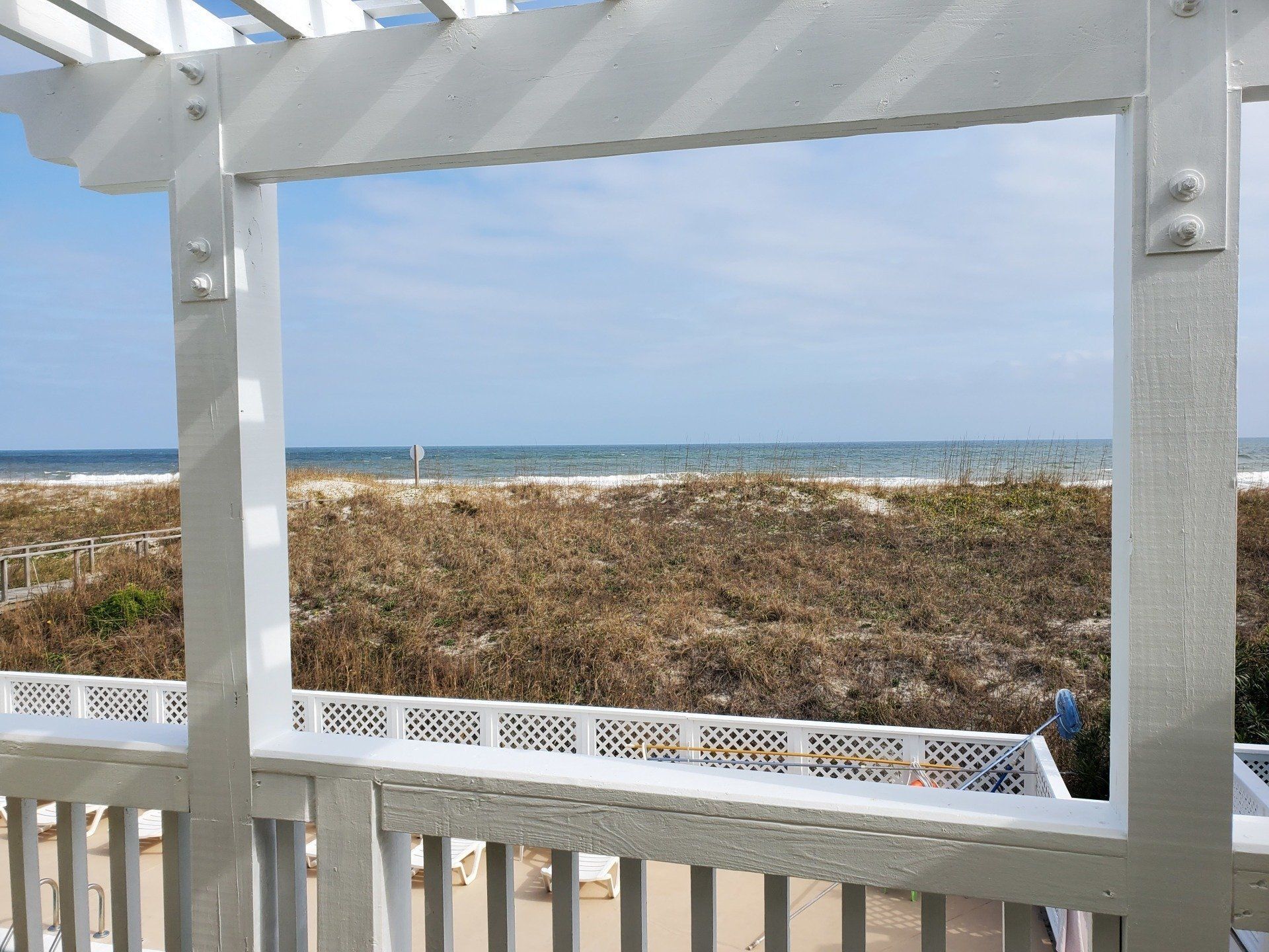 A view of the ocean and sand dunes framed by a white wooden pergola and railing on a sunny day.