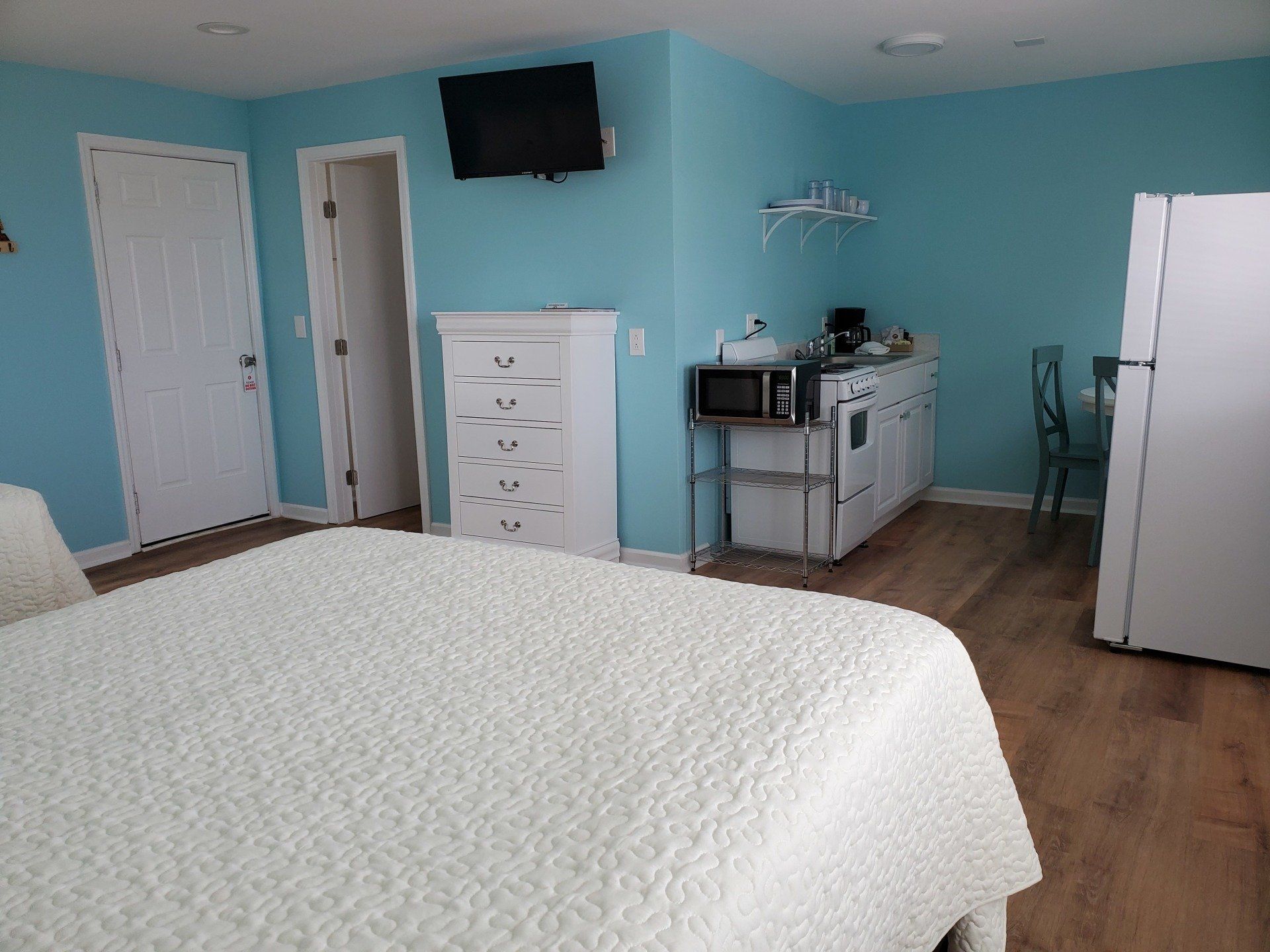 A hotel room with light blue walls, a white bed, a white dresser, a wall-mounted TV, a kitchenette, and a refrigerator.