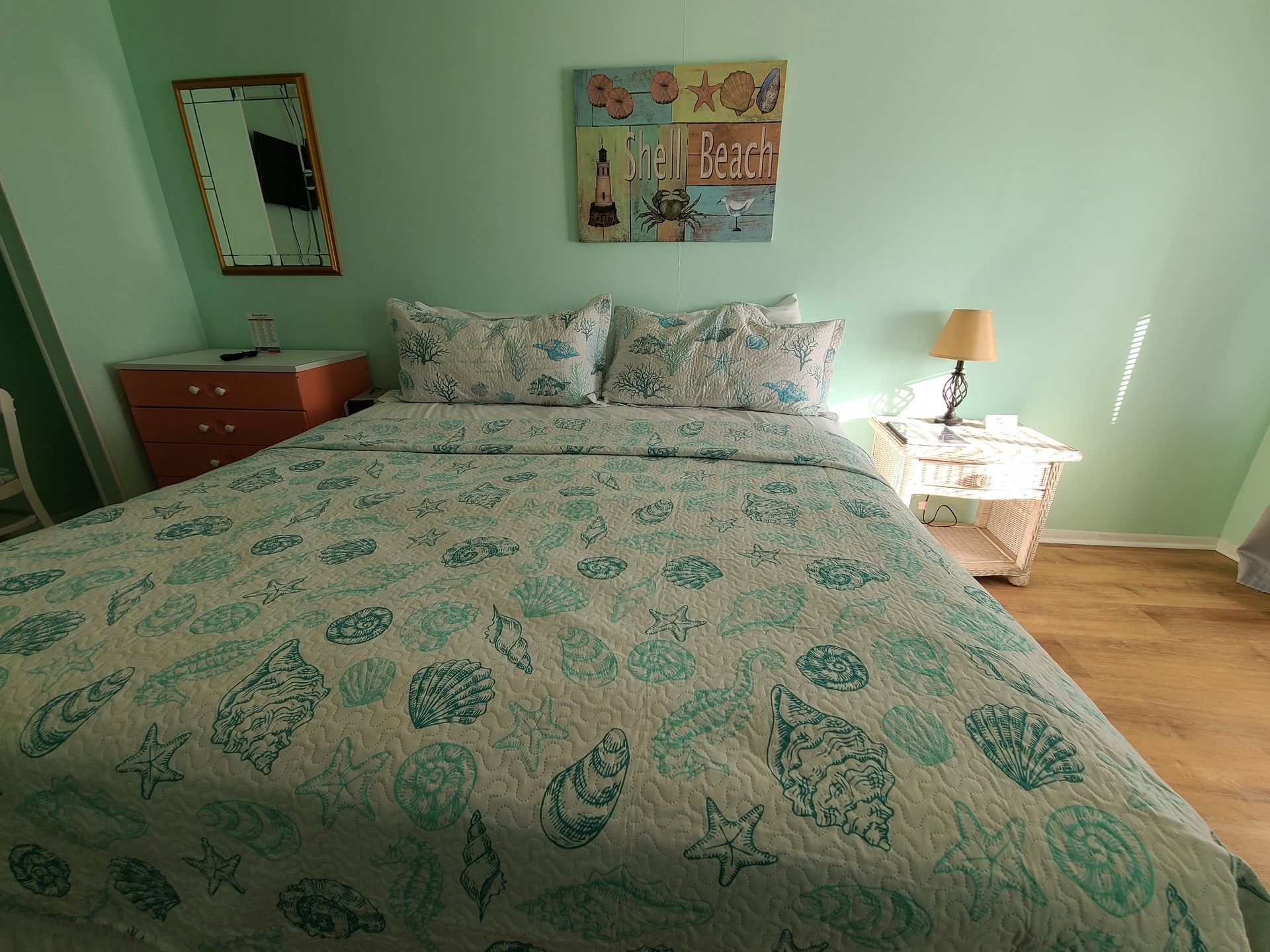 A bedroom with a bed featuring a teal seashell-patterned quilt, a nightstand with a lamp, and a wall mirror.