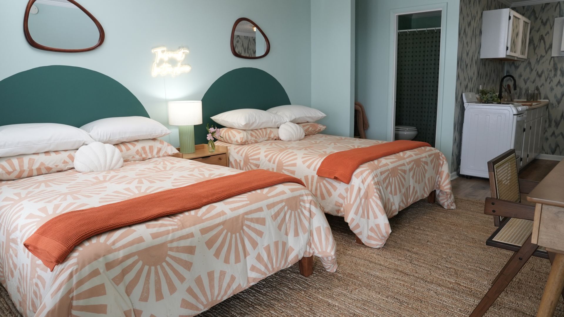 A bedroom with two queen beds, teal headboards, patterned bedding, orange throws, and a small kitchen area in the back.