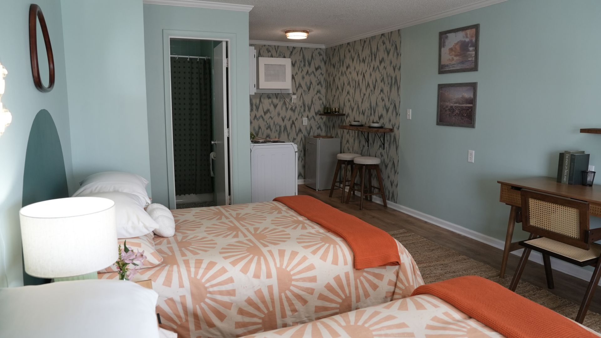 A hotel room with two beds, a small desk, two stools, and a corner kitchenette with light blue walls and patterned paper.
