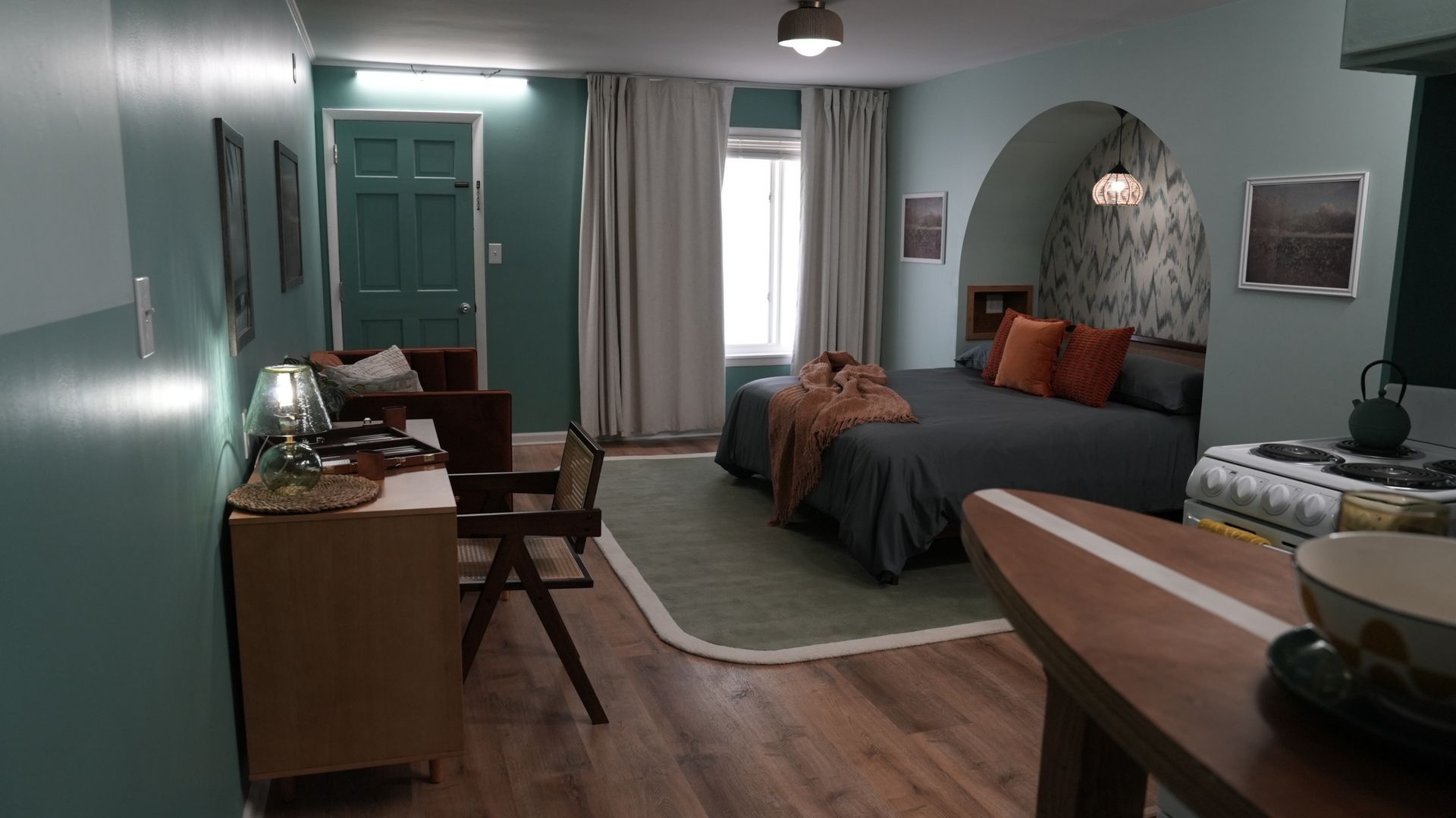 A bedroom with a teal wall, a bed set in an arched alcove with orange pillows, light curtains, and wooden flooring.