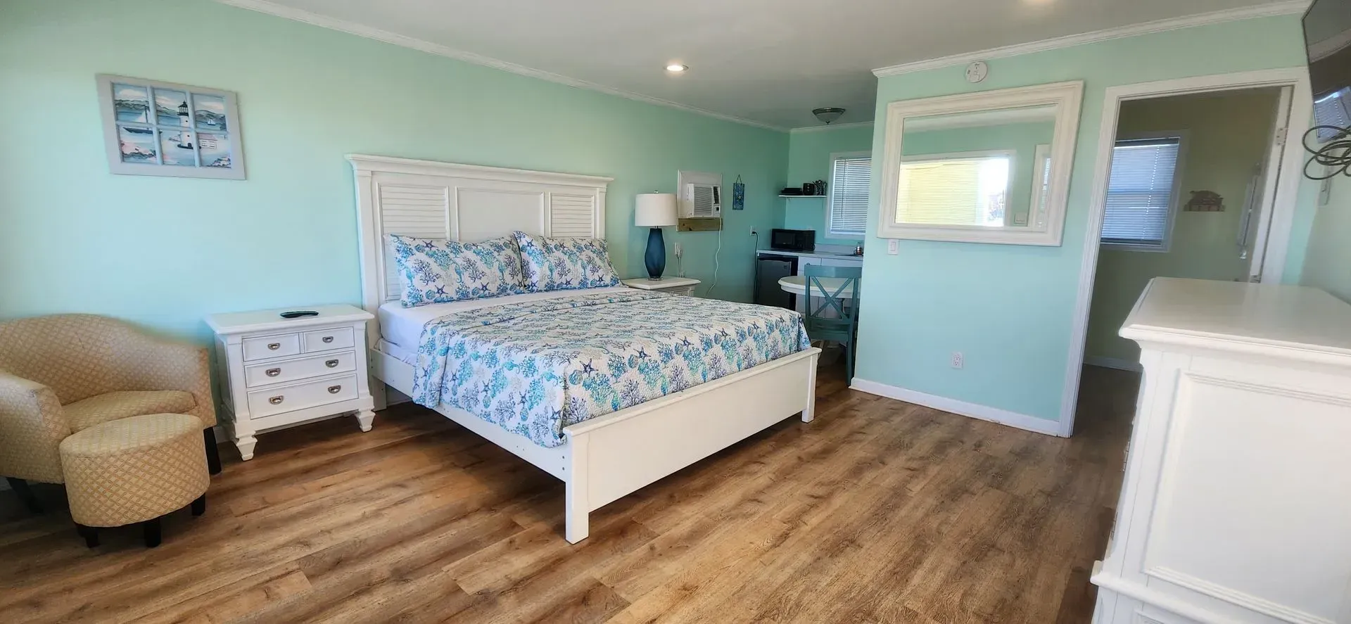 A bright bedroom with a bed featuring blue and white patterned bedding, a cream armchair, and white furniture on wood floors.