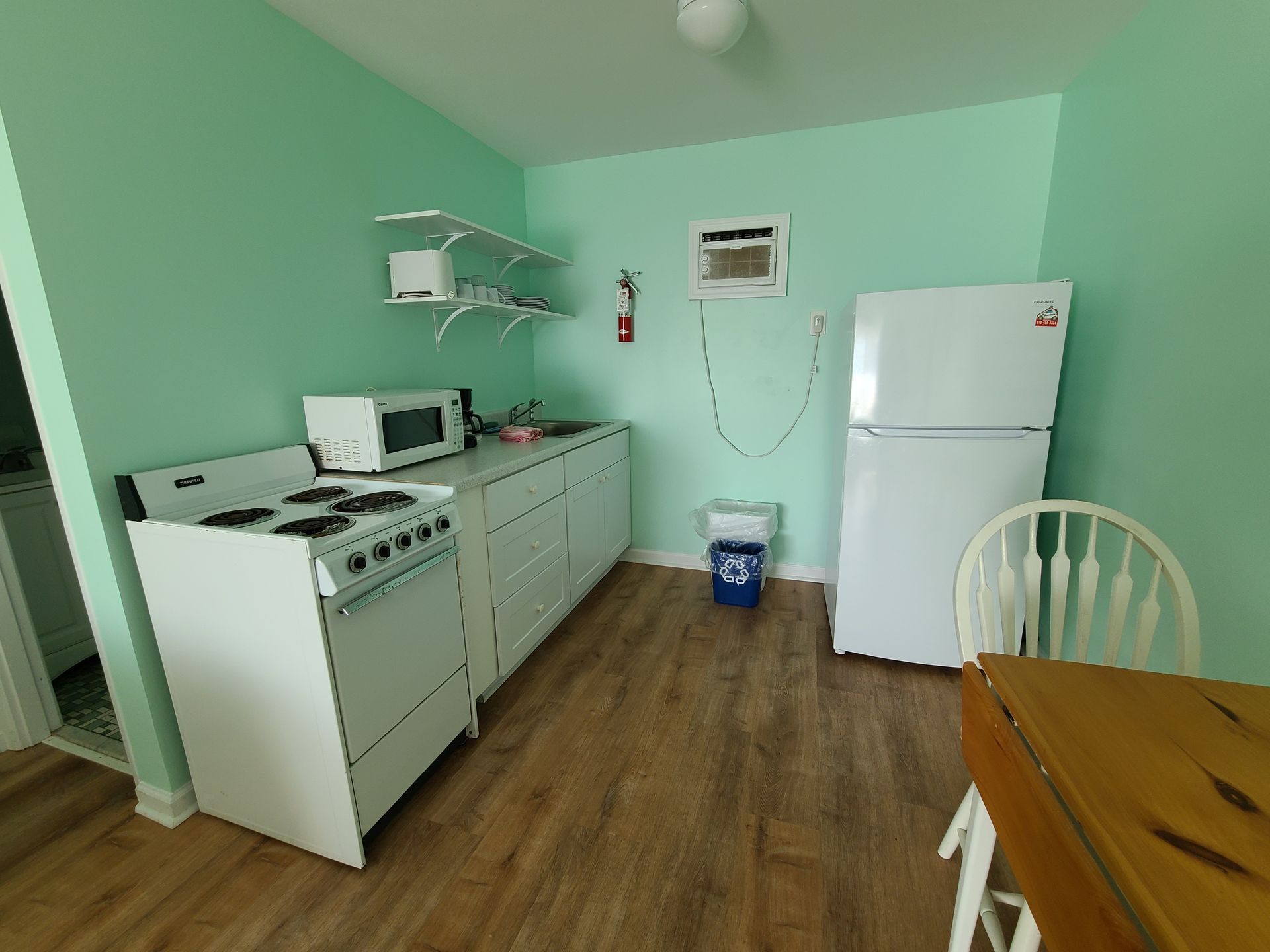 A small kitchen with light green walls, white appliances, wooden flooring, a stove, a refrigerator, and a dining chair.