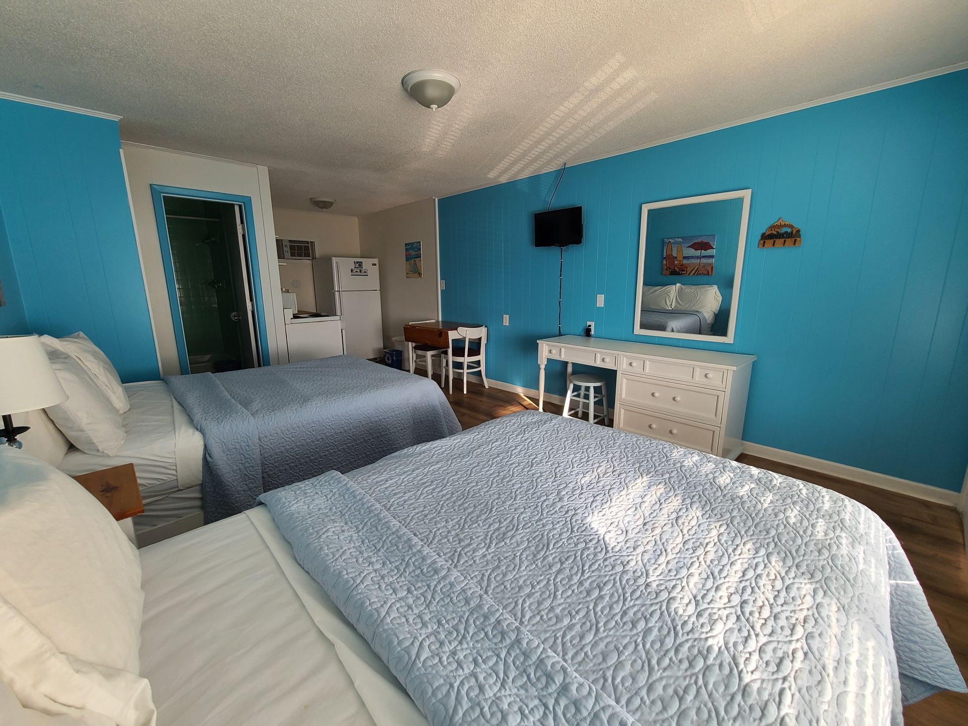 A hotel room with two queen beds, bright blue walls, a white vanity, a mirror, and a small refrigerator.