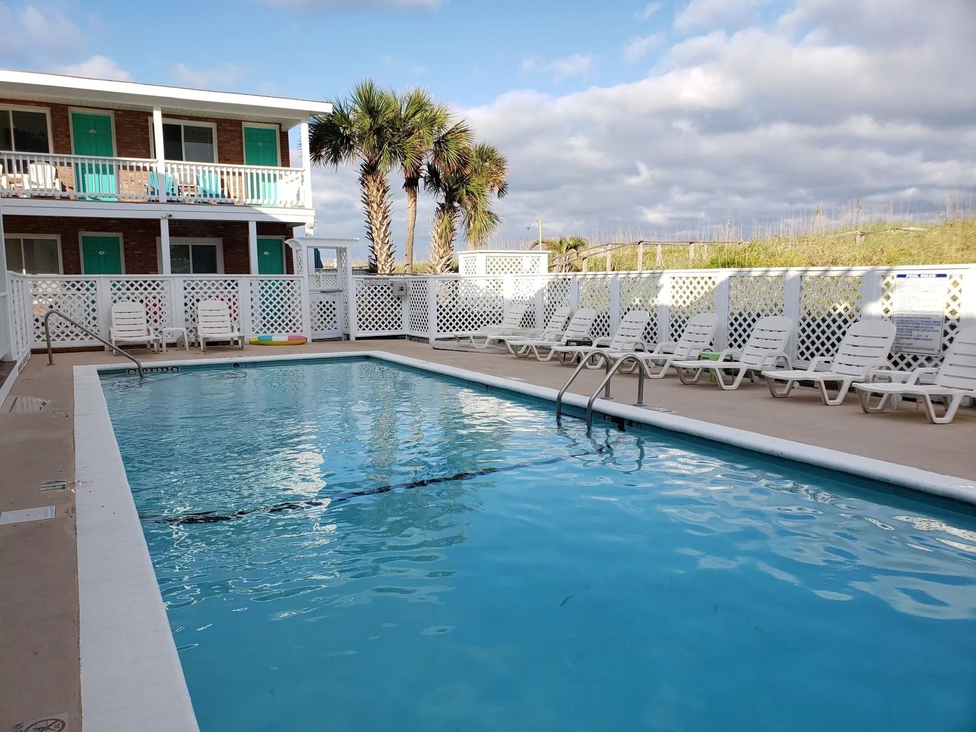 A rectangular swimming pool with blue water sits in front of a two-story motel with white railings and turquoise doors.