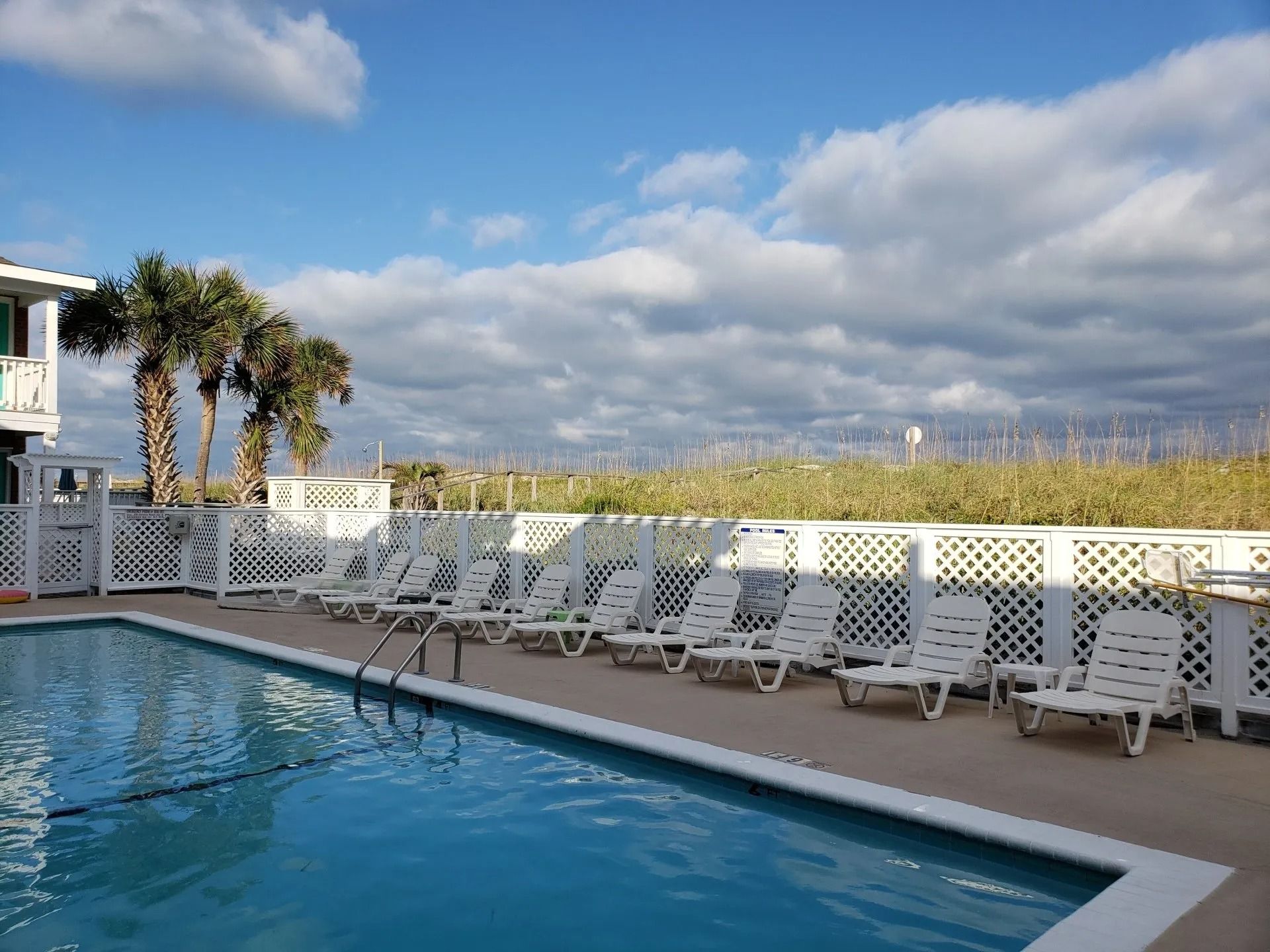 A swimming pool with lounge chairs on a deck, bordered by a white lattice fence, palm trees, and a cloudy blue sky.