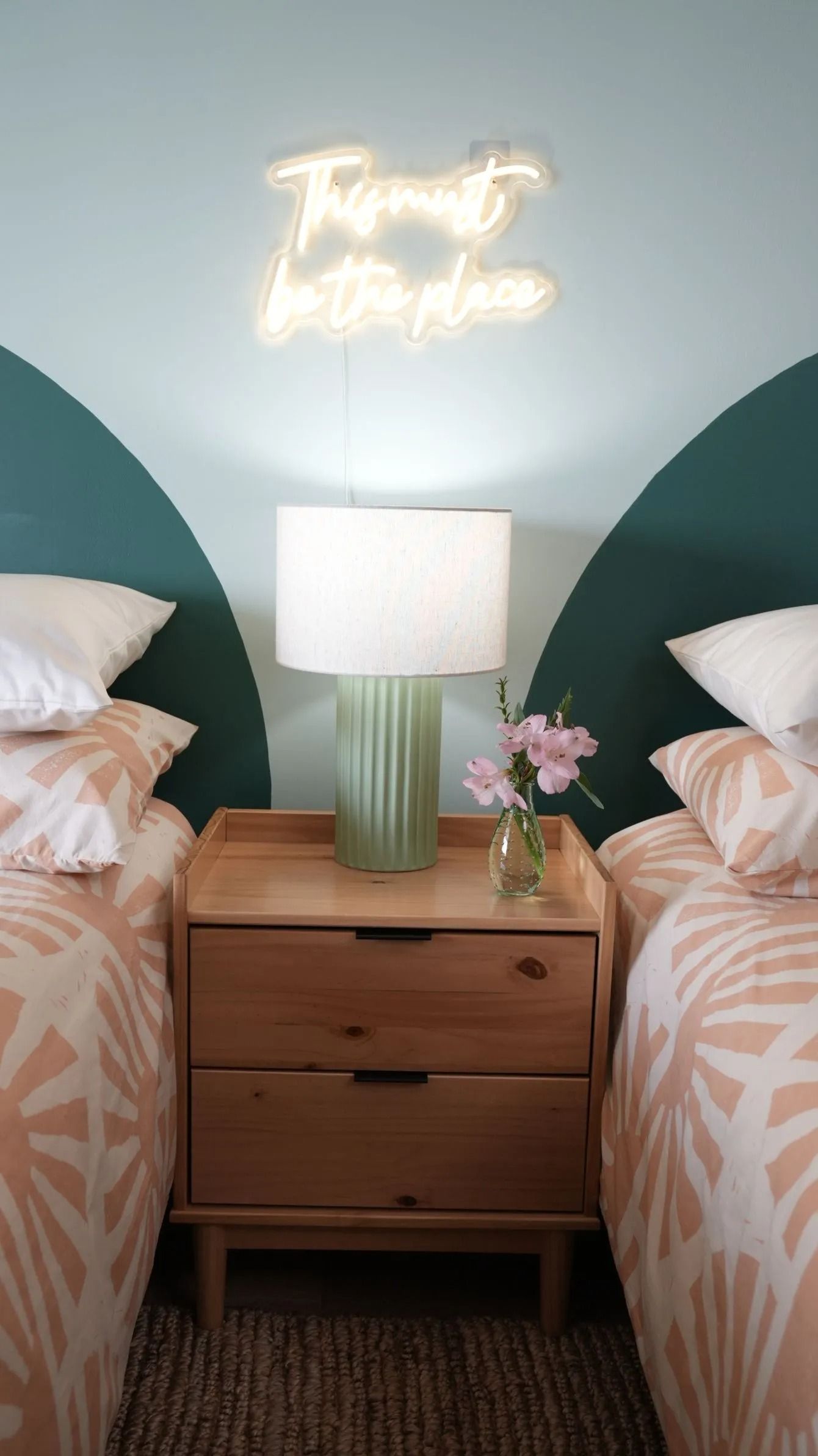 A wooden nightstand with a lamp and vase sits between two beds against a wall with two painted dark green circular arcs.