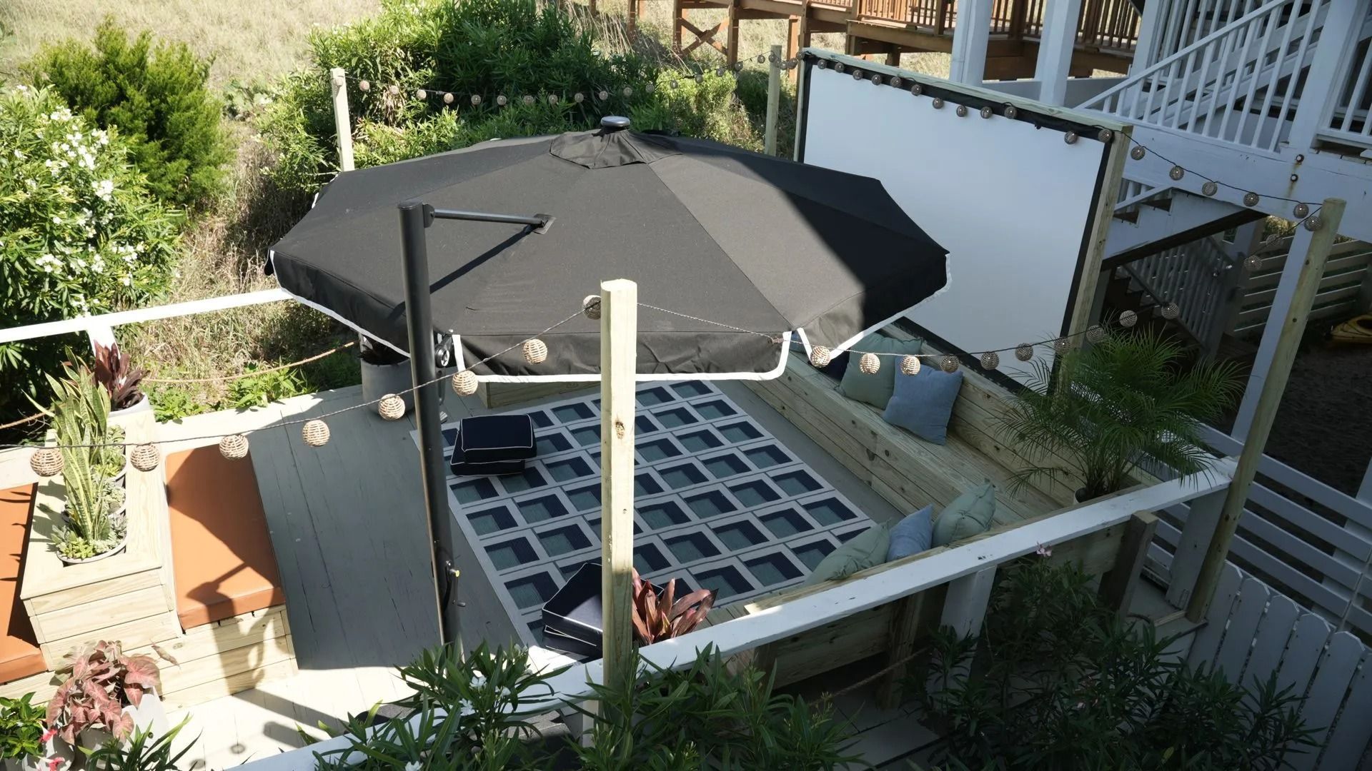 A high-angle view of a backyard patio with a grid-patterned floor, a black umbrella, seating, and a large white screen.