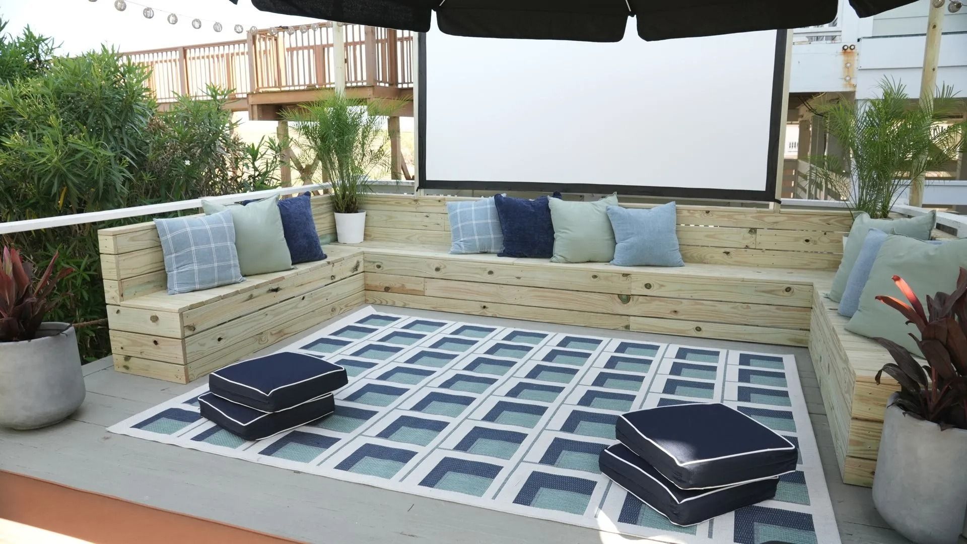 An outdoor lounge with wooden bench seating, blue cushions, a projection screen, and a geometric-patterned rug on a patio.