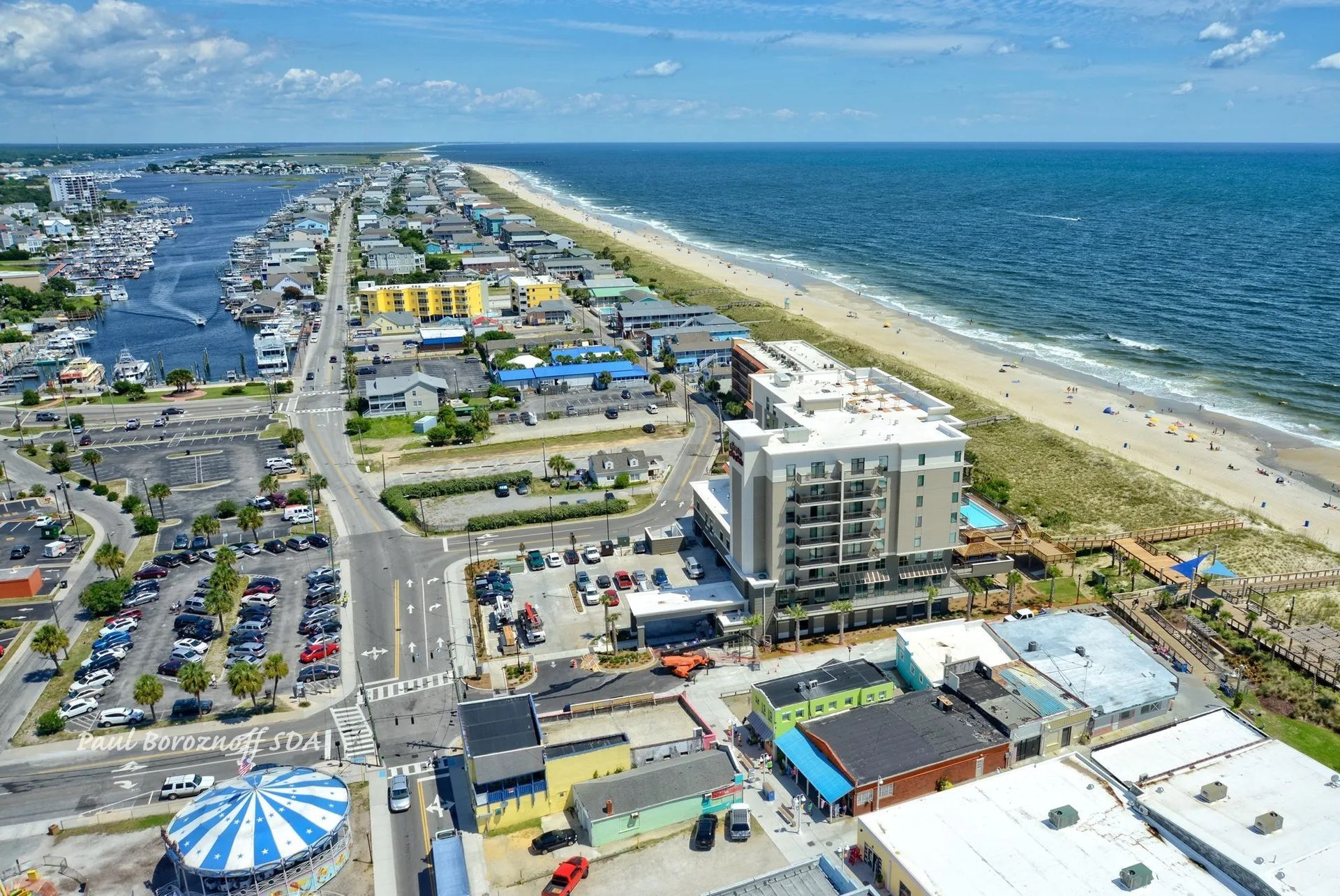 An aerial view of a coastal beach town with hotels, parking lots, and a harbor, beside the ocean under a blue sky.