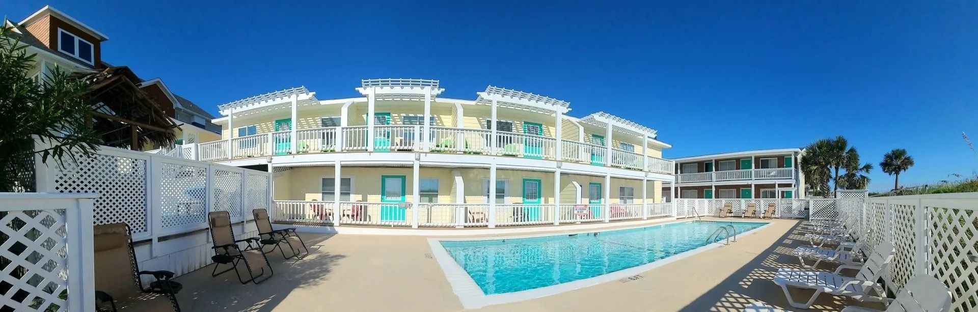 A panoramic view of a sunny resort featuring a bright blue swimming pool, lounge chairs, and two-story cream-colored units.
