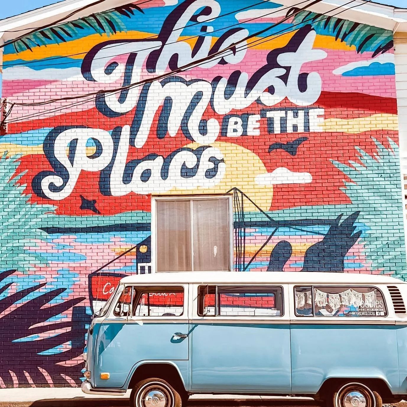 A light blue vintage Volkswagen bus parked in front of a colorful brick wall mural that says,